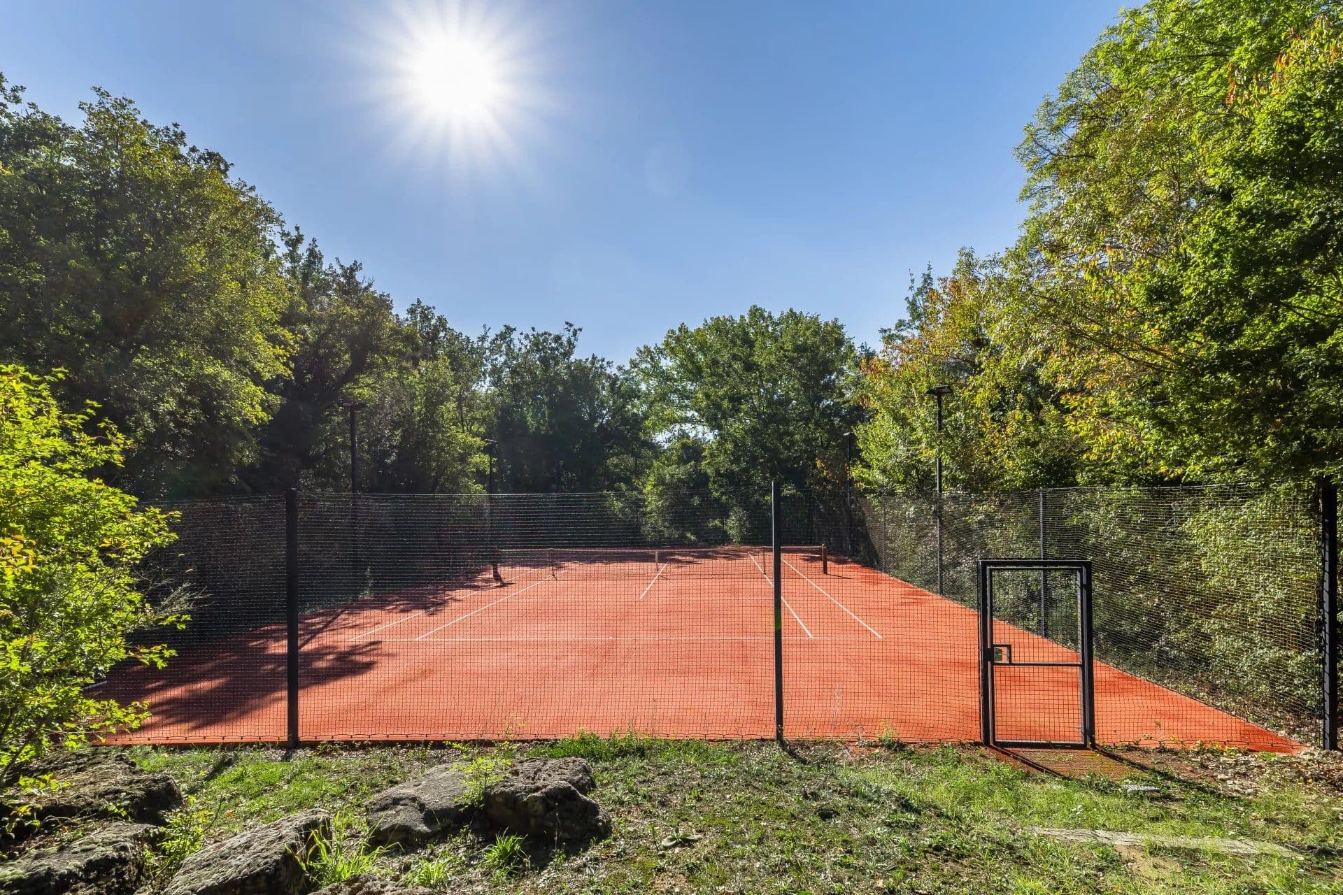 Un court de tennis extérieur rouge entouré d'arbres verts sous un ciel lumineux et ensoleillé. La zone est entourée d'une clôture noire, avec une petite porte visible.