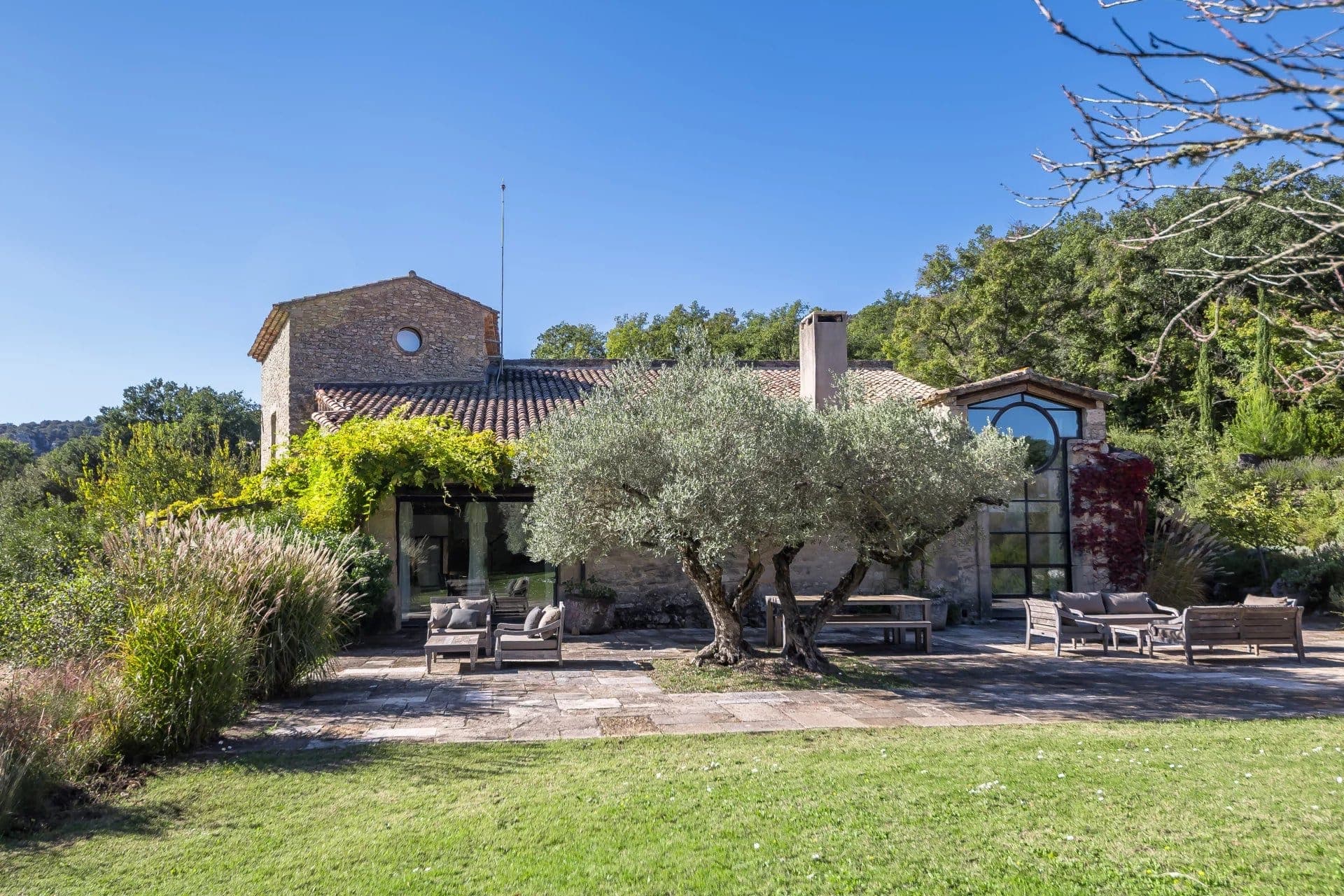 Villa en pierre avec toit en tuiles, située dans un jardin luxuriant. Grandes fenêtres, terrasse avec mobilier de jardin et deux oliviers. Entourée de verdure et sous un ciel bleu clair, elle dégage une atmosphère paisible et rustique.
