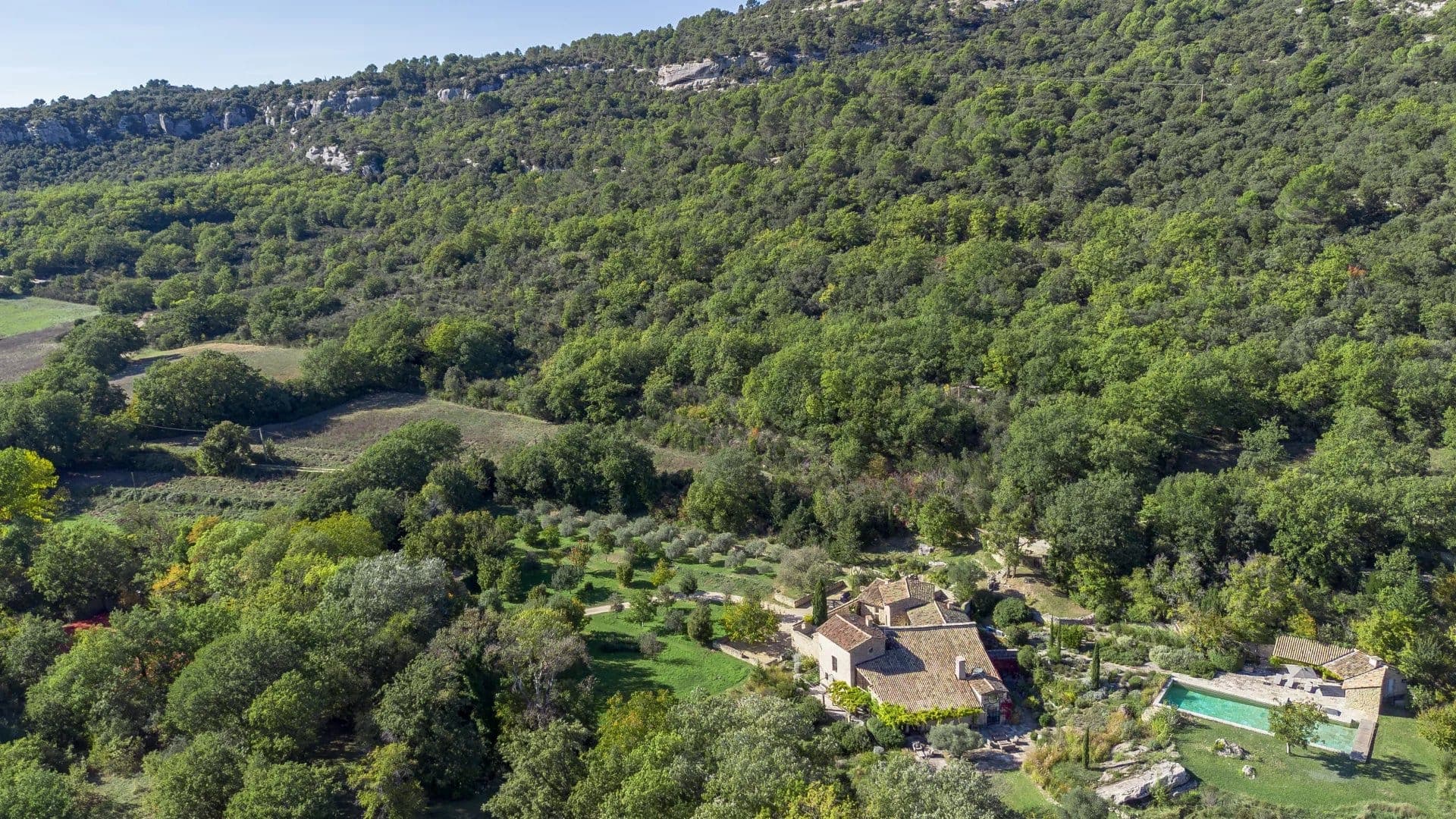 Aerial view of a lush, hilly landscape with a rustic house and a swimming pool surrounded by greenery. The area is dense with trees and shrubs, and the terrain is elevated with a clear blue sky above.