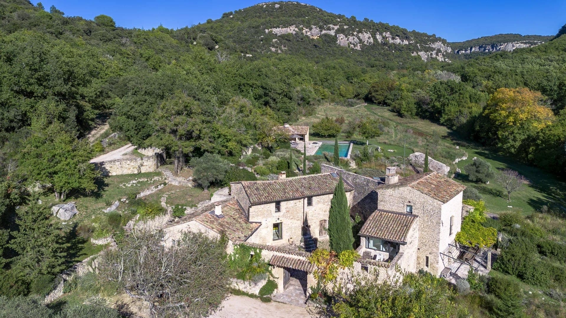Aerial view of a rustic stone house nestled in a lush, green landscape with hills in the background. The house features multiple pitched roofs, a courtyard, and nearby verdant gardens. A hidden swimming pool is partially visible among the trees.