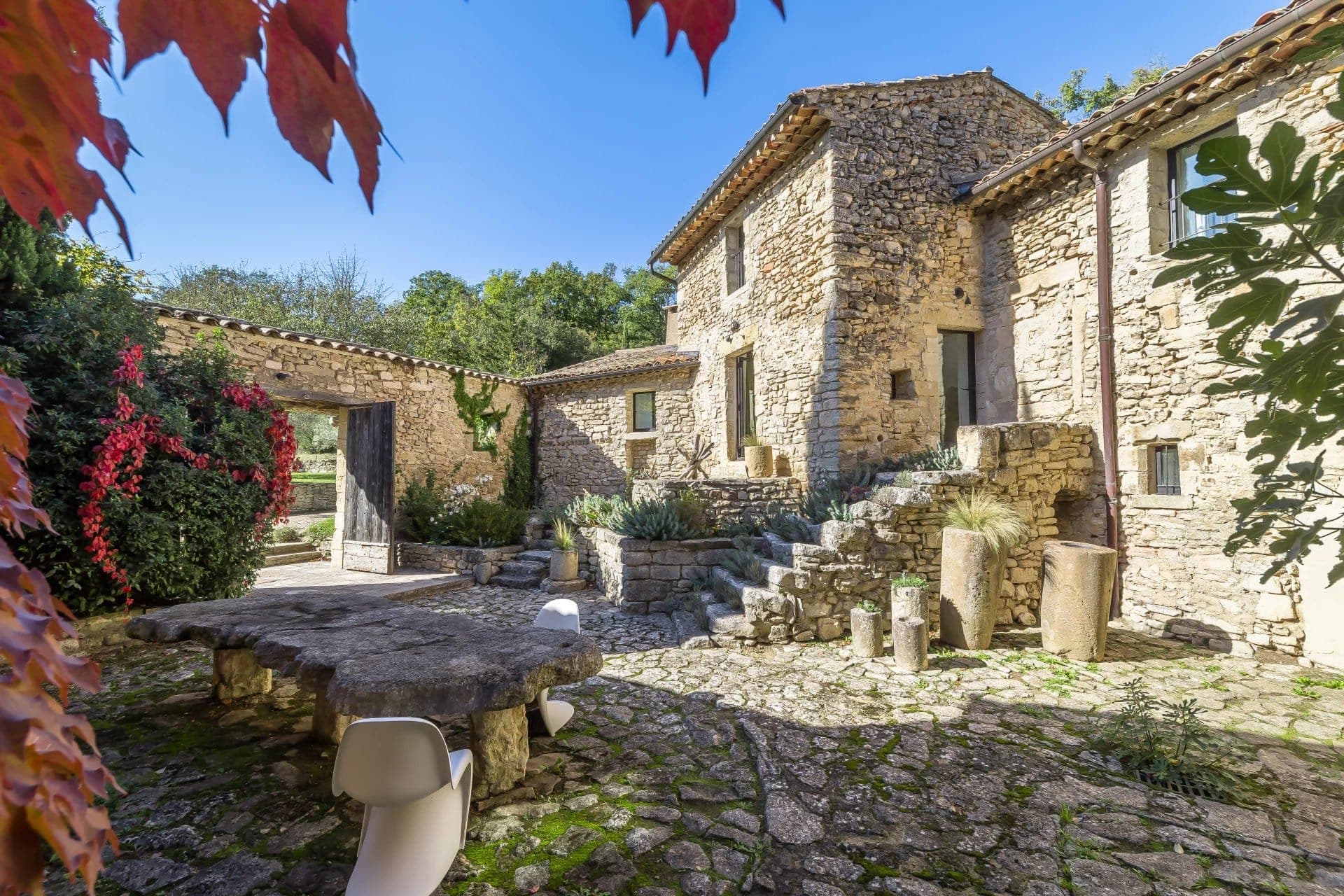 A rustic stone courtyard with a weathered table and modern chairs. Stone buildings surround the area, with steps leading to a door. Red leaves frame the edge, and greenery is visible around the courtyard under a clear blue sky.