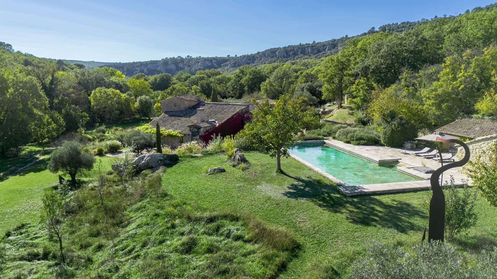 A serene landscape shows a lush green yard with a swimming pool, surrounded by trees and stone sidewalks. In the background, there's a house with a red wall, nestled among more trees and rolling hills. The sky is clear and blue.