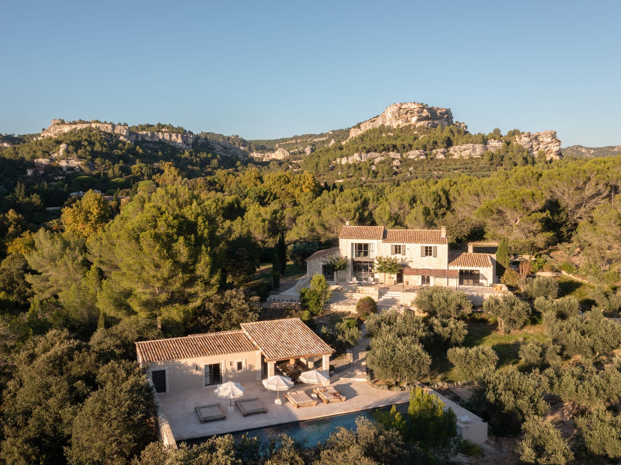 Aerial view of a luxury hillside villa surrounded by lush trees and rocky cliffs under a clear blue sky. The property features a pool, sun loungers, and multiple buildings with terracotta roofs and stone walls, set in a picturesque landscape.