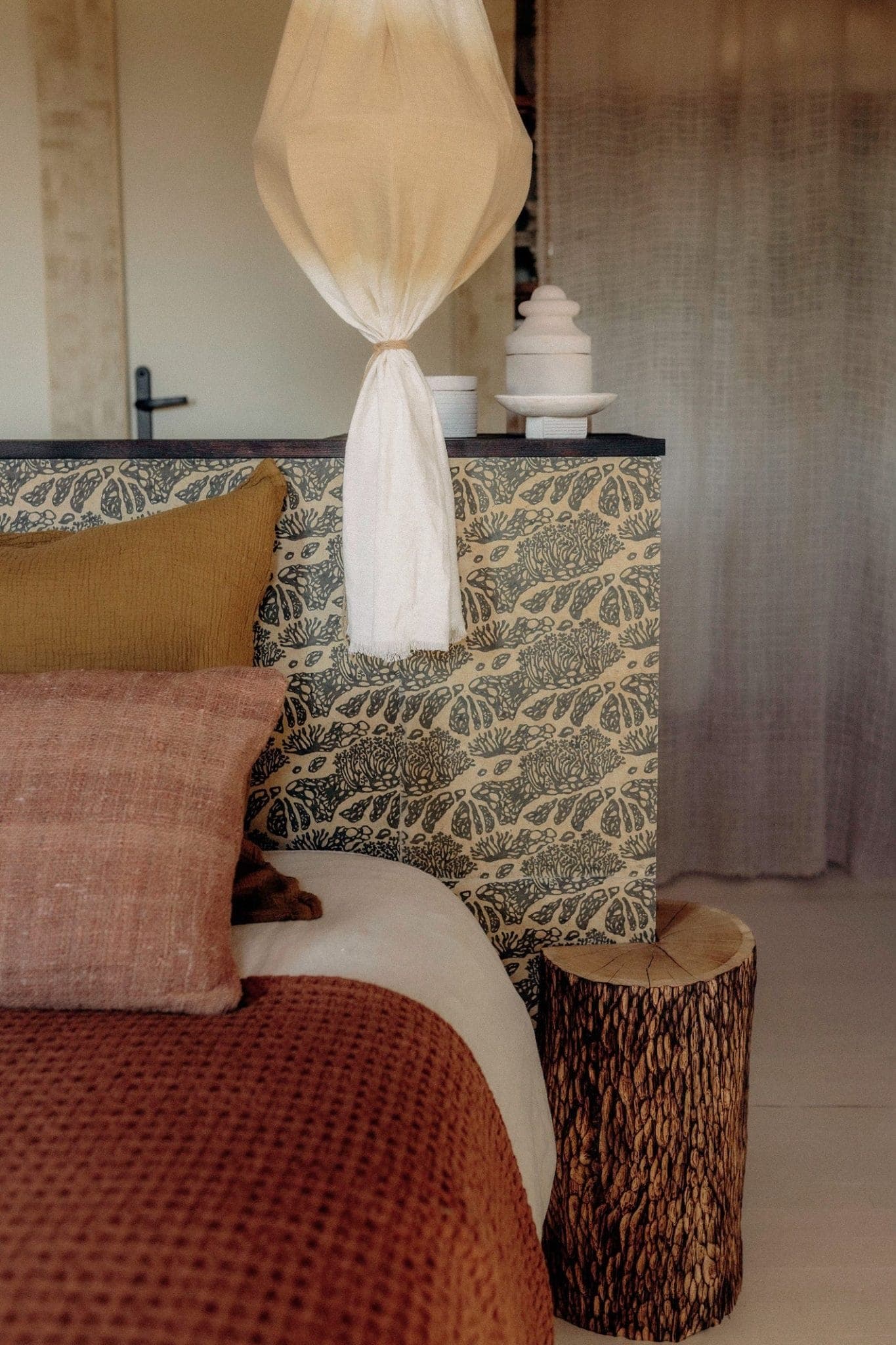 Cozy bedroom with a bed featuring brown and beige pillows and a textured bedspread. A rustic wood nightstand sits beside the bed. The headboard has a leafy pattern, and a hanging fabric lamp adds a soft, warm glow to the room.