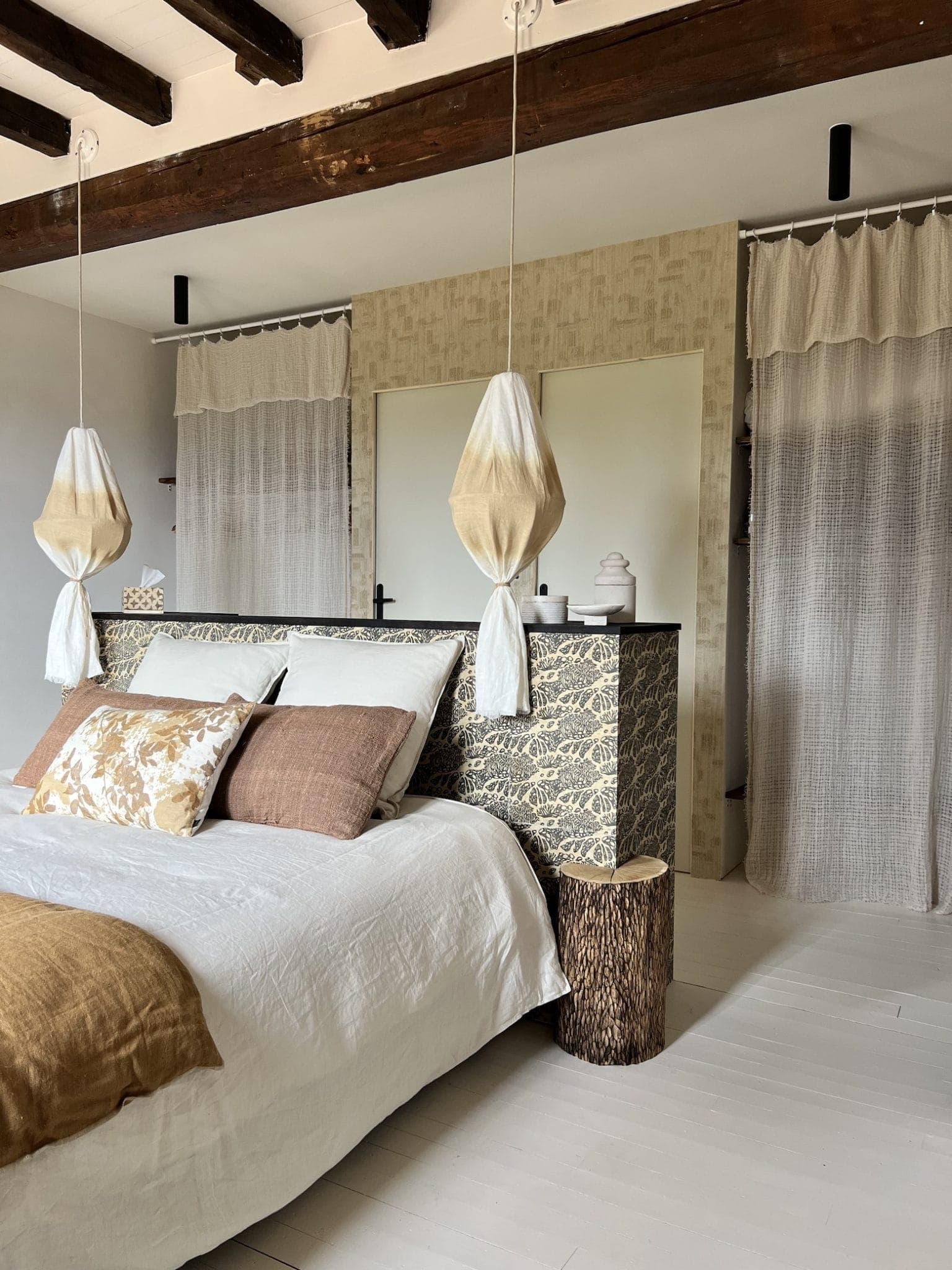 A cozy bedroom with a bed featuring white and brown pillows. Above the bed, two decorative pendant lights hang. The room has rustic wooden beams, a patterned headboard, and sheer curtains separating a dressing area in the background.