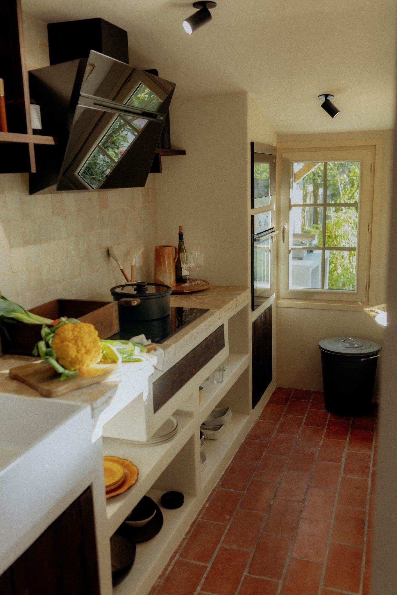 A cozy, narrow kitchen with terracotta floor tiles, light walls, and a window. It features dark wooden cabinets, a stove with a pot, and fresh vegetables like cauliflower and lettuce on the countertop. A black trash bin is near the window.