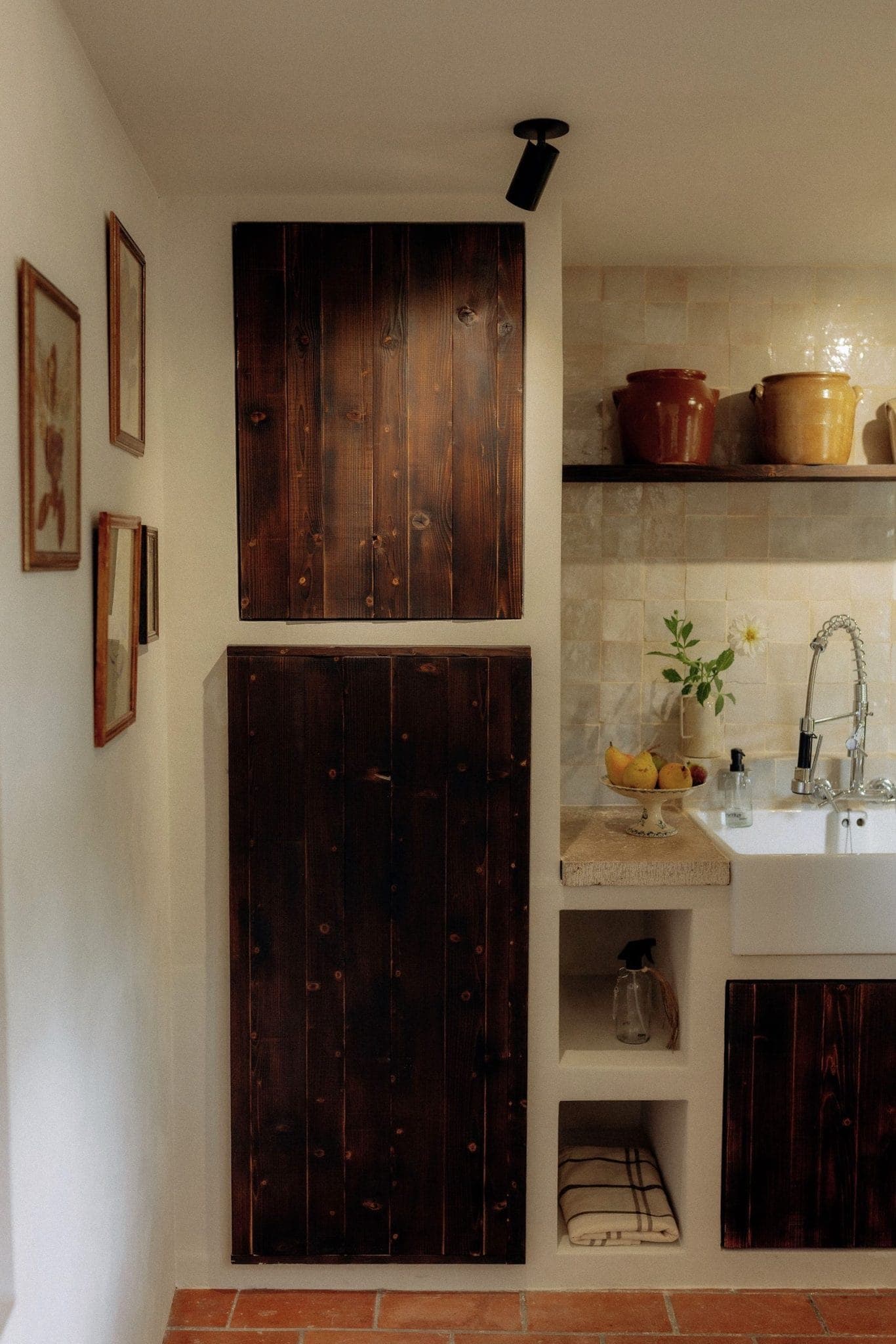 Cozy kitchen with dark wooden cabinets, beige tile backsplash, and rustic decor. A white sink sits next to potted plants and a basket of lemons on the counter. Framed art hangs on the wall, and terracotta floor tiles complete the warm ambiance.