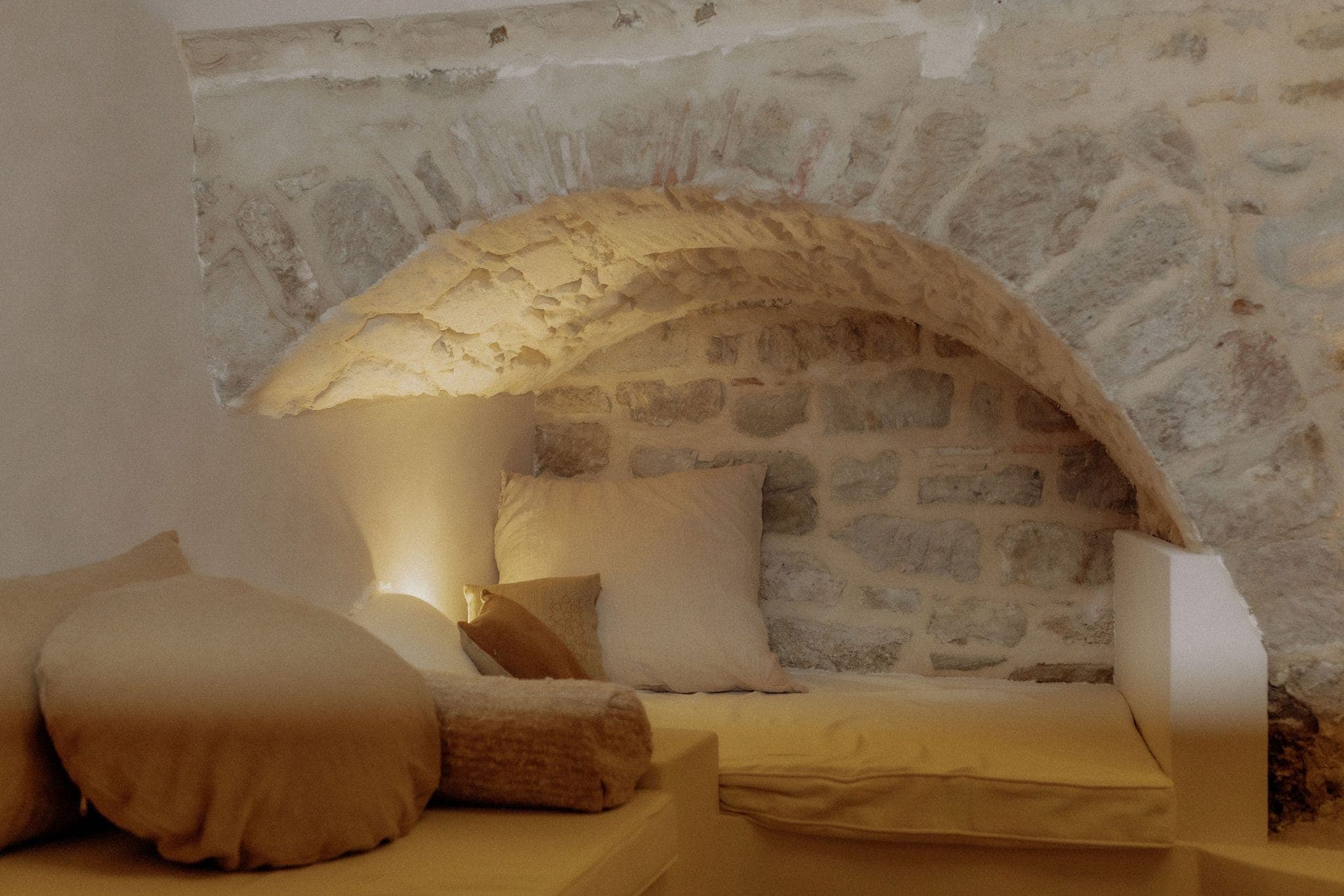 A cozy nook with a stone wall and an arched ceiling, featuring plush beige cushions on a low, cushioned seating area. Soft lighting creates a warm ambiance, highlighting the textured stone and white walls.
