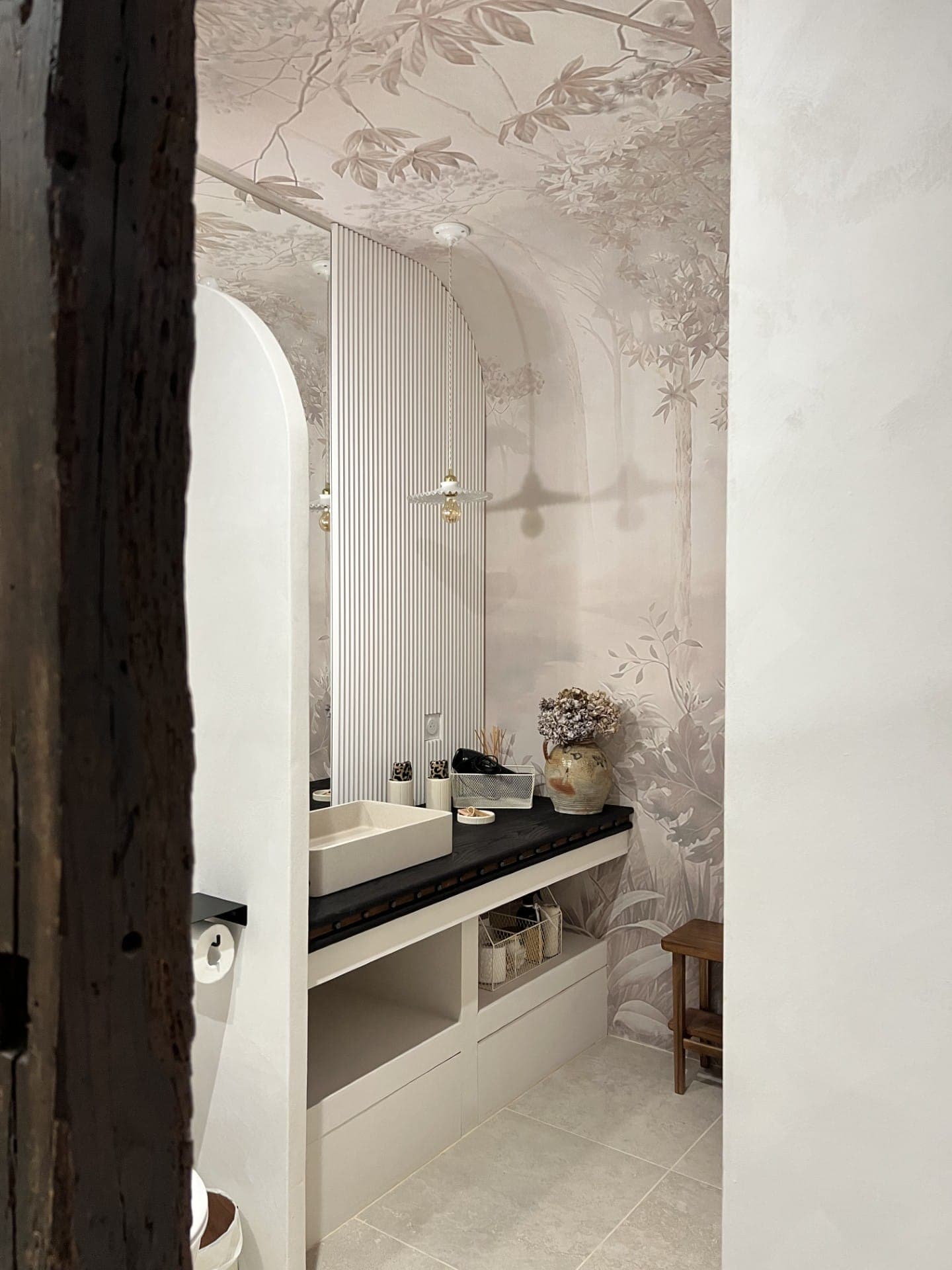 Elegant bathroom with a large mirror and a white sink on a black countertop. The walls feature a nature-themed mural. There's a potted plant and decorative vases. A wooden stool sits to the side, and soft lighting creates a welcoming atmosphere.