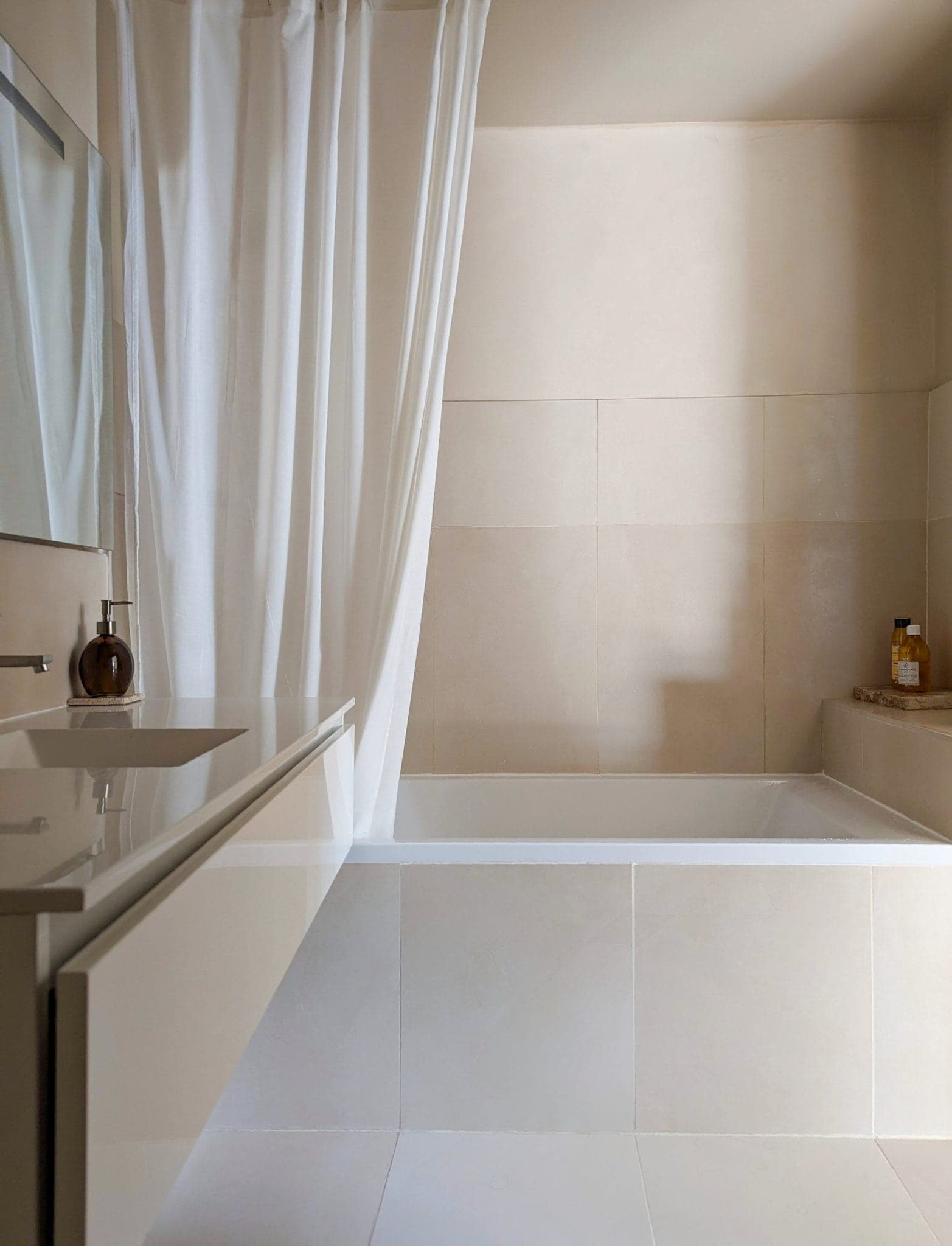Minimalist bathroom with white bathtub and curtain. Beige tiles cover the walls. A white sink with a dark soap dispenser is on the left. A small bottle and container sit on the edge of the tub. A large mirror hangs over the sink.