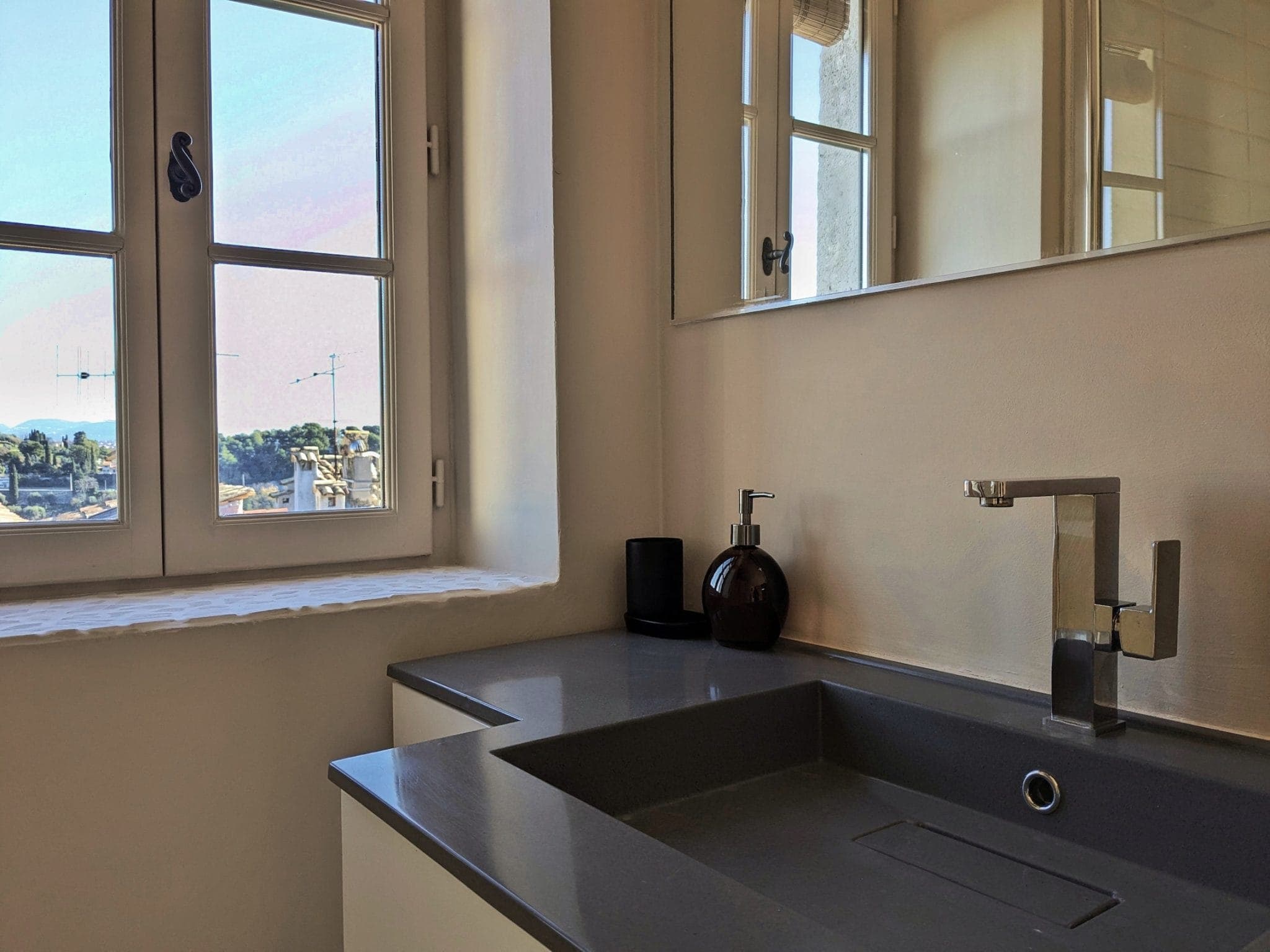 A modern bathroom with a sleek gray countertop and sink. A silver faucet sits next to a round, dark soap dispenser. A large window offers a view of the trees and outdoor buildings, adding natural light to the space.