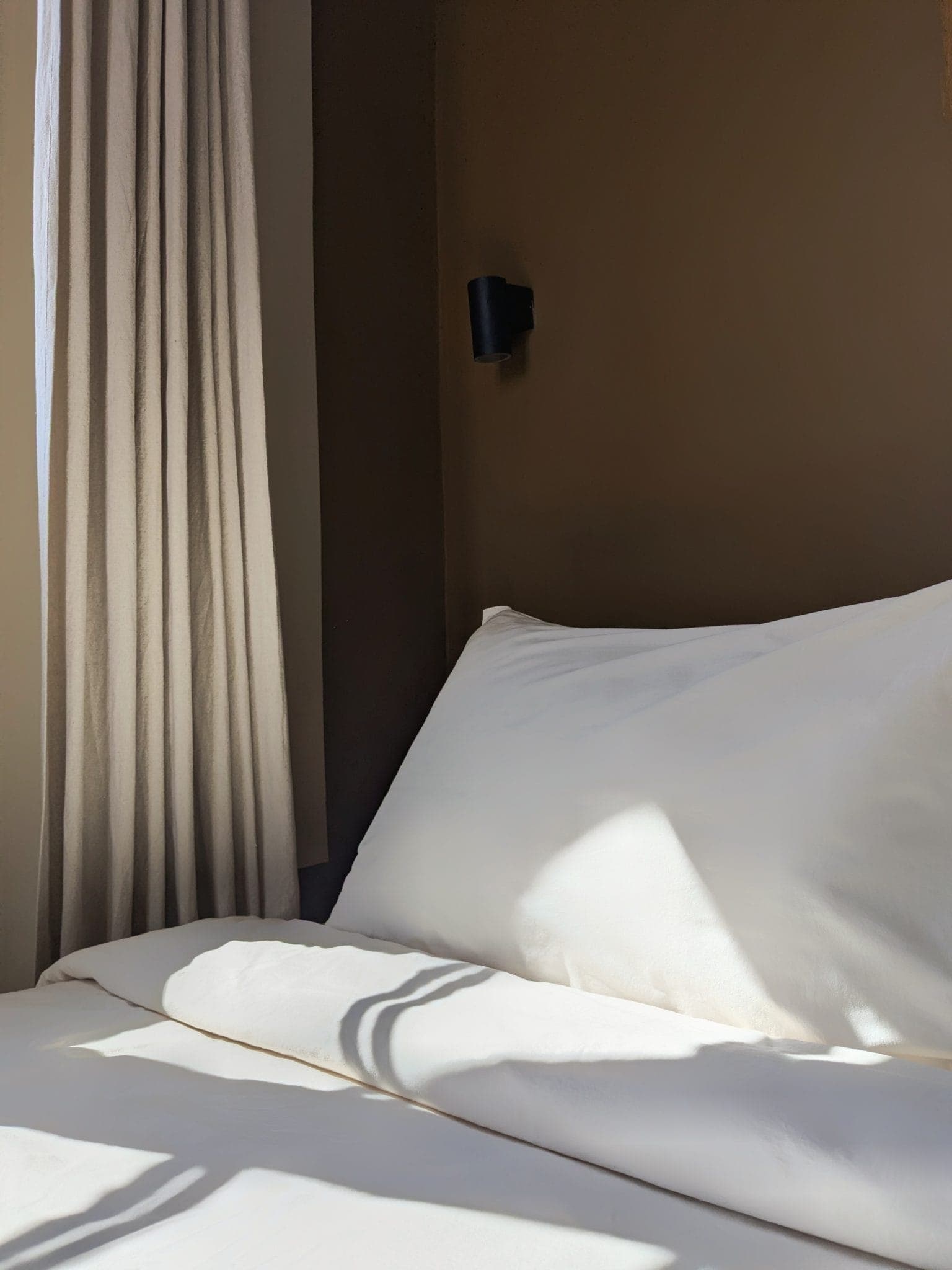 A well-made bed, with white sheets and pillows, is bathed in sunlight. A beige curtain hangs next to a brown wall, to which a small black light fixture is attached. Shadows cast on the bed add depth to the scene.