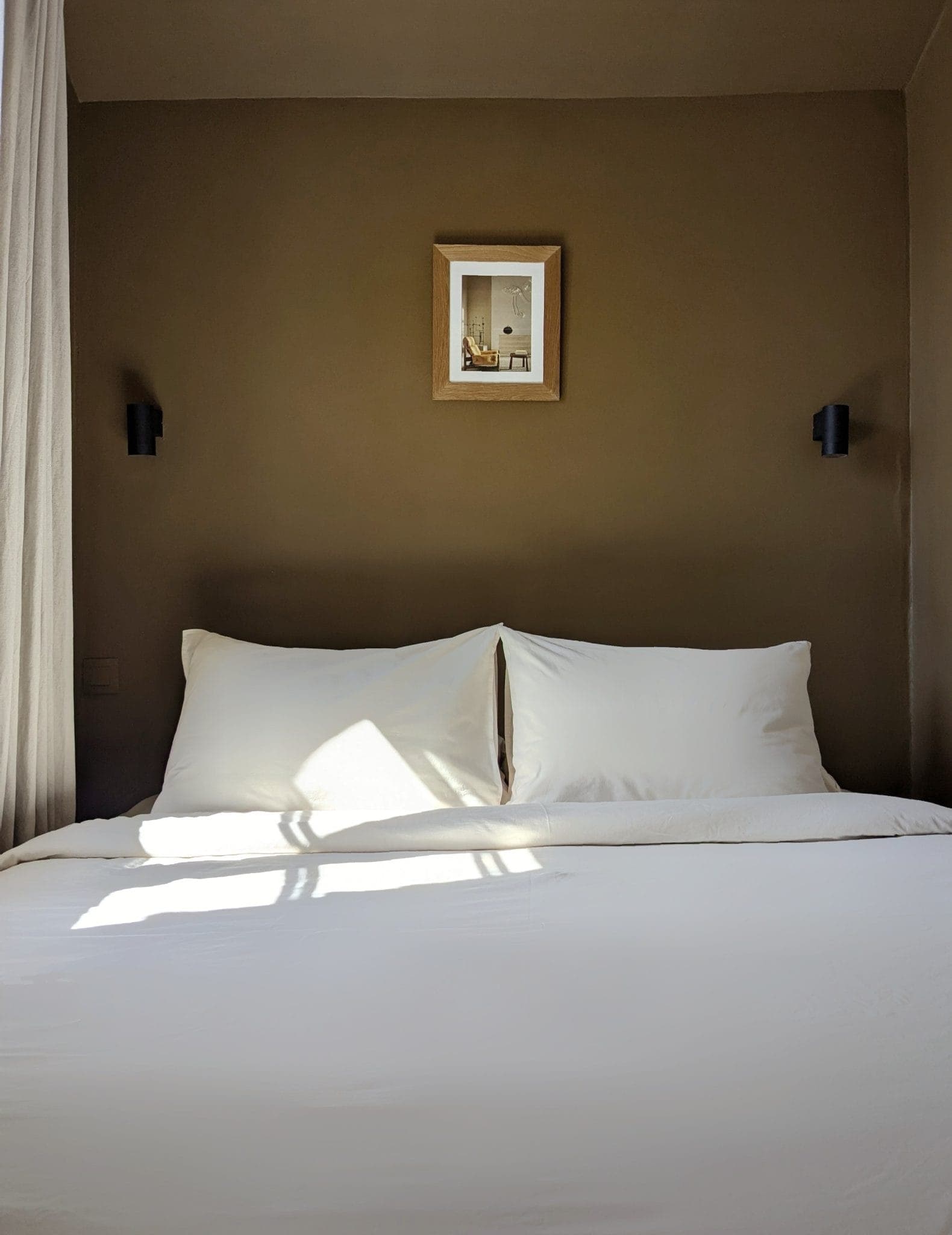 A well-made bed with white sheets is set against an olive-green wall. Above the bed is a small picture frame. Natural light casts shadows on the bed from a nearby window. Two black wall lamps frame the bed.