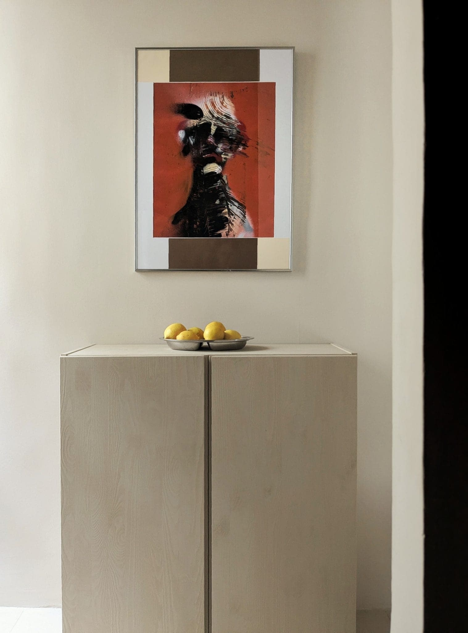 A minimalist interior with a light-wood cabinet topped by a bowl of yellow lemons. Above the cabinet, a framed abstract painting with bold, dark strokes on a bright red background is displayed on a light-colored wall.