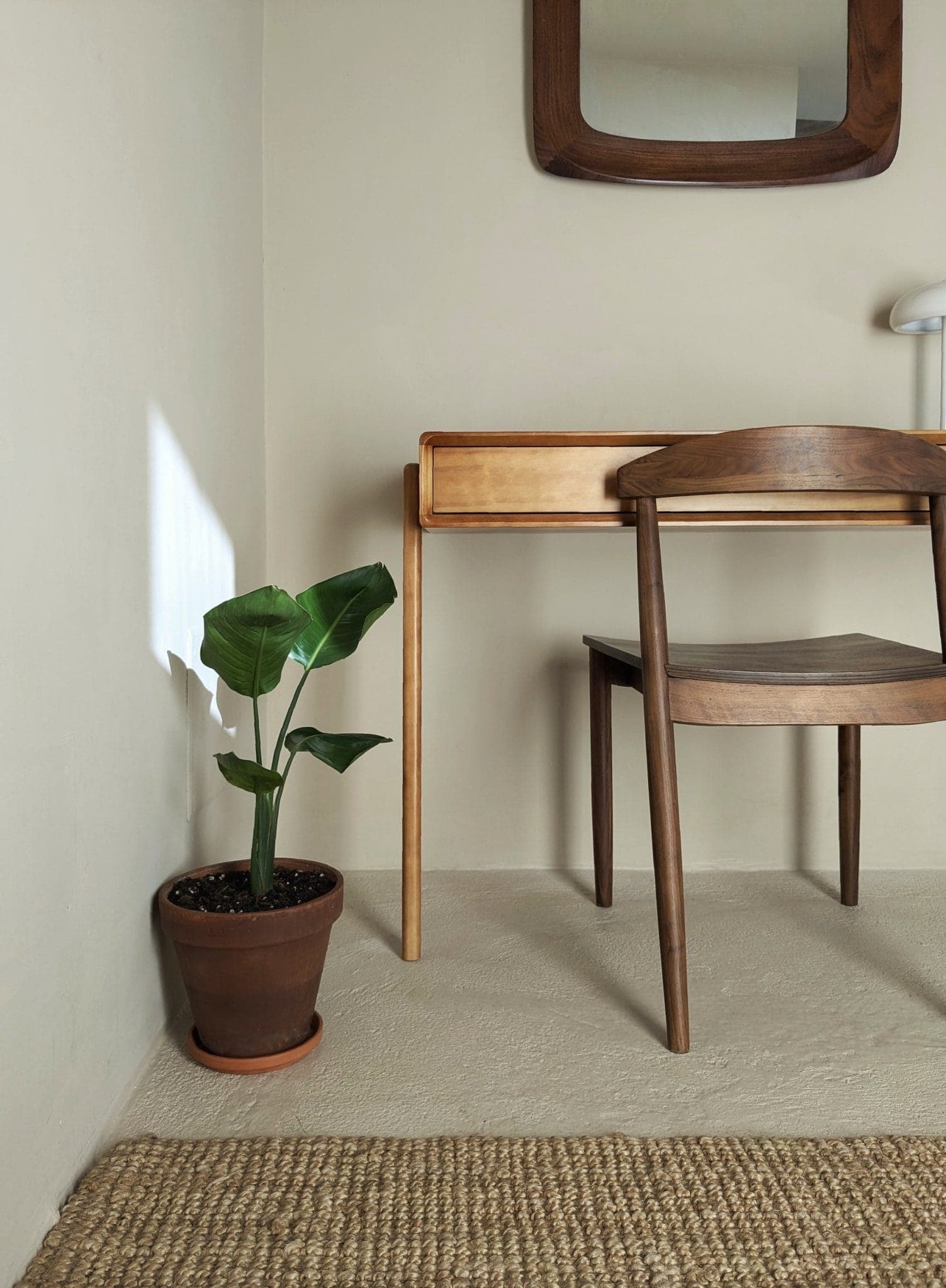 A minimalist workspace features a wooden desk and chair against a light beige wall. A rectangular mirror hangs above the desk. A small potted plant sits on the floor next to a woven rug, while sunlight casts a shadow on the wall.