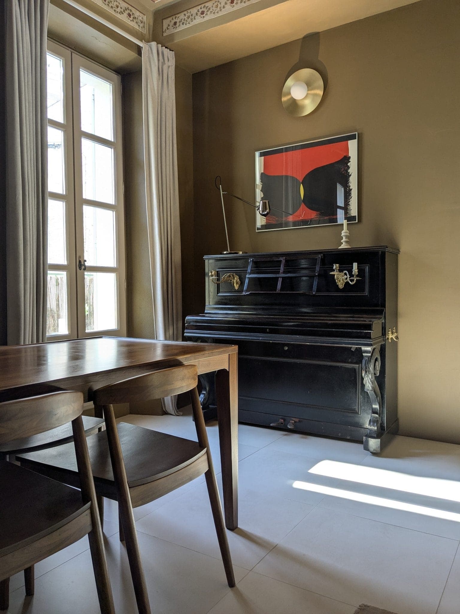 A dining area with a wooden table and chairs next to a black upright piano. Above the piano is a black and red abstract work of art. Natural light streams in through a window with beige curtains. A circular wall lamp sits above the painting.