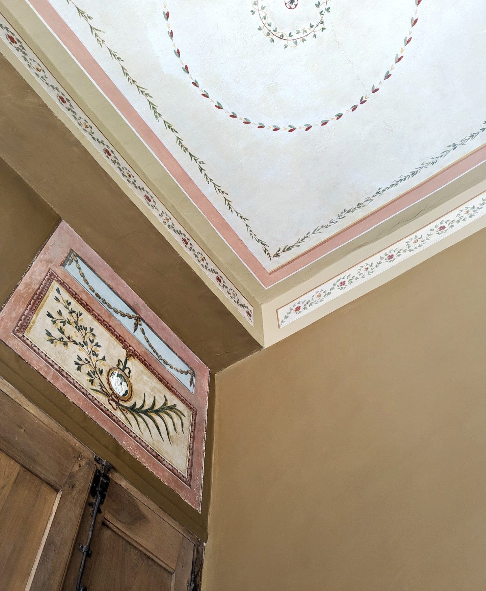The picture shows a corner of a room with floral wall and ceiling decorations. The ceiling features circular floral motifs, while the wall has a rectangular floral painting above the wooden doors. The colors are neutral tones of beige, pink and green.