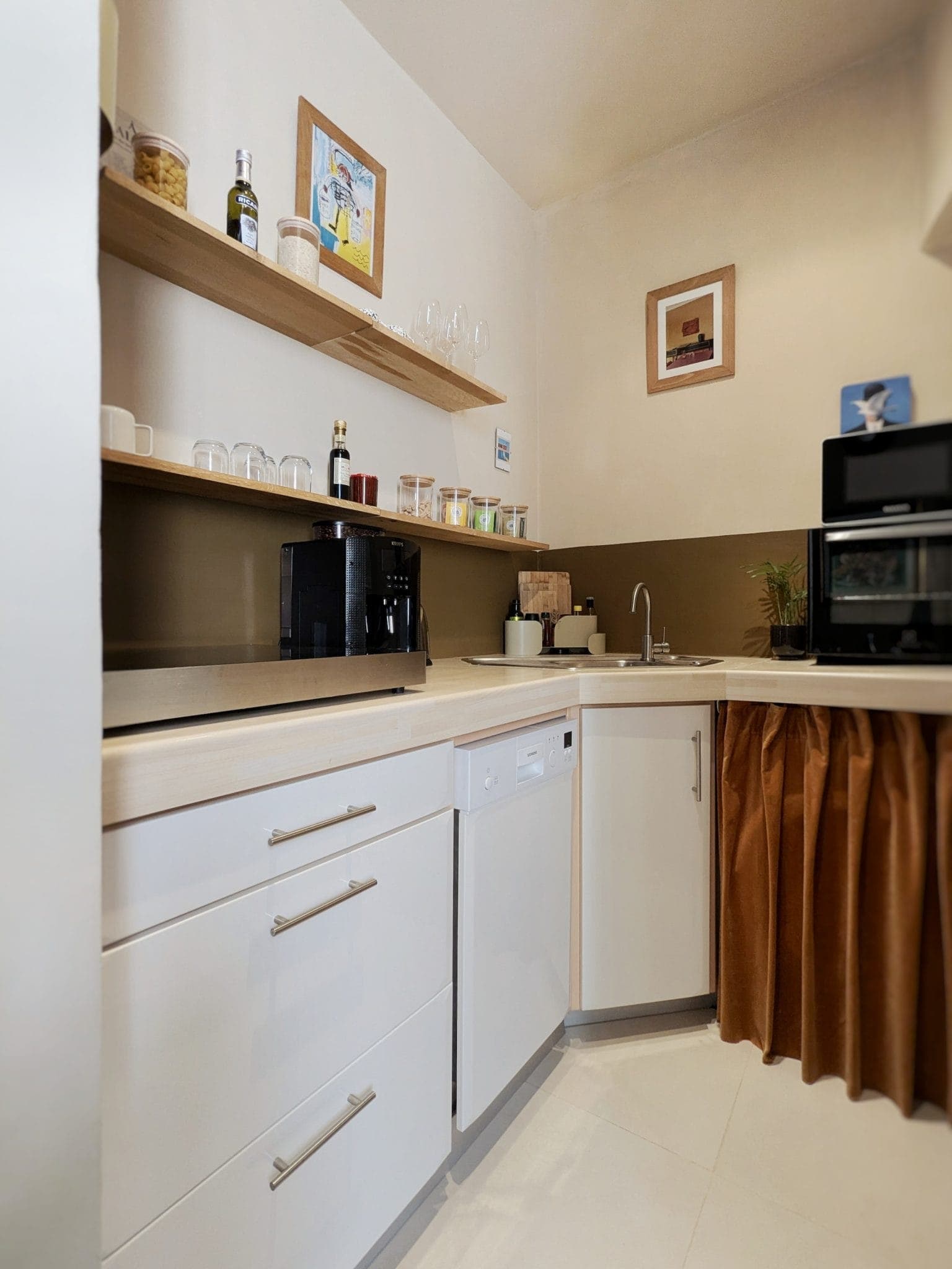 Modern kitchenette with white cabinets and a brown curtain under the sink. Open shelves hold glasses, cups and bottles. A coffee machine sits on the counter and a built-in oven is visible. Framed works of art hang on the walls.