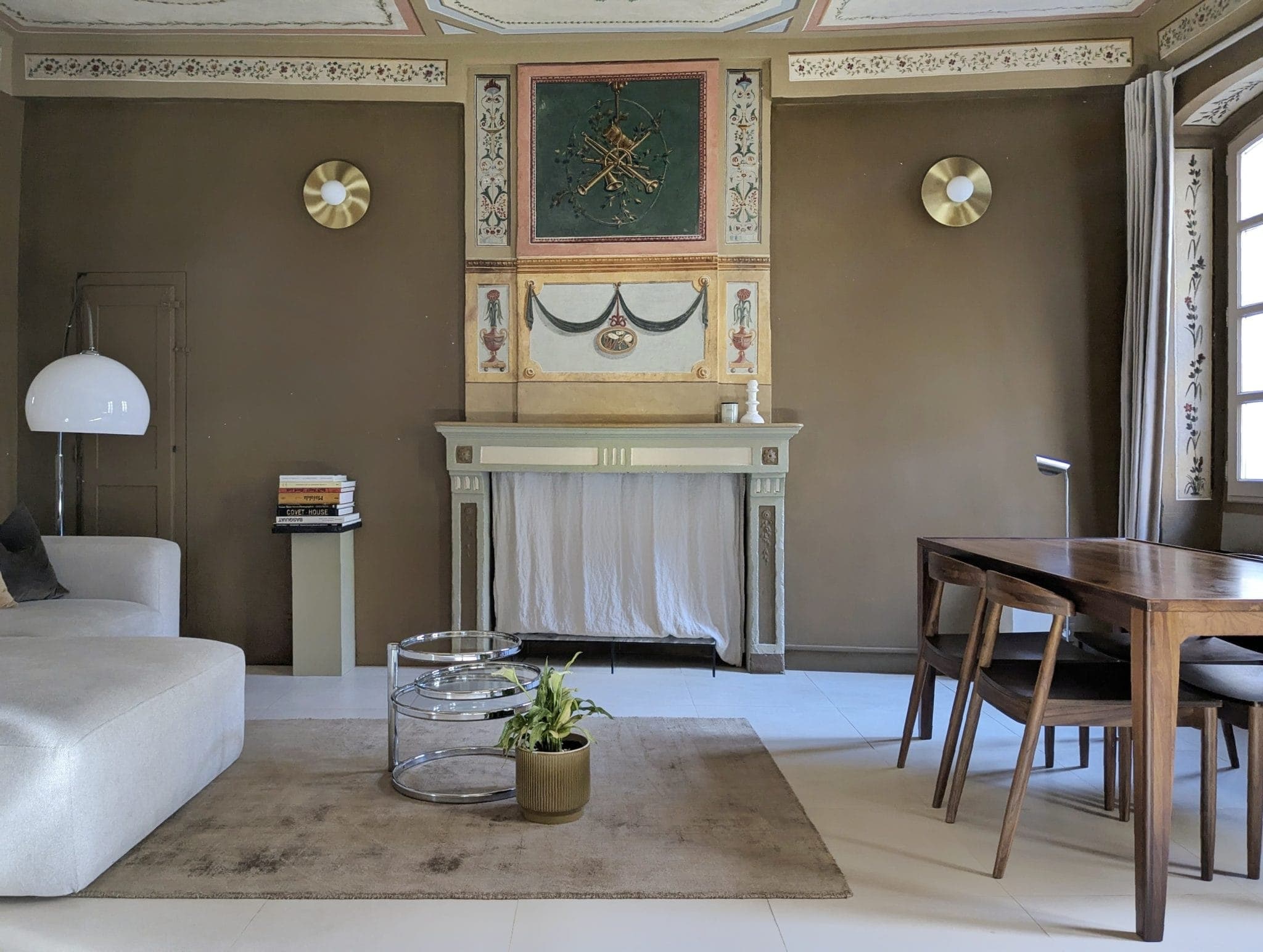 A stylish living room with brown walls features an ornate fireplace, a white lamp, a beige couch, a wooden dining table with chairs, and a round glass coffee table on a beige rug with a small potted plant on it. Sunlight streams through large windows.