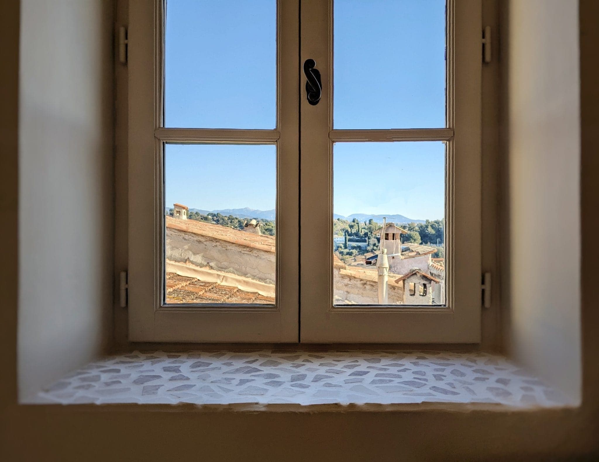 A white-framed window overlooks a small village with tiled roofs and a distant landscape of hills under a clear blue sky. The windowsill is covered with a mosaic pattern.