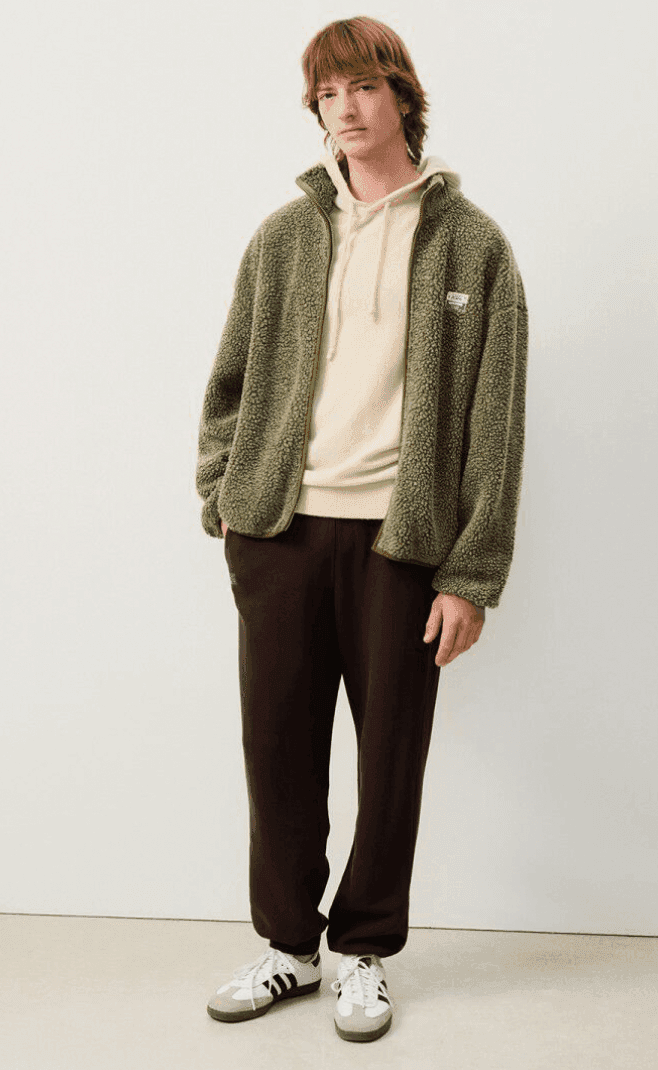 A person with medium-length hair stands against a plain background wearing a green fleece jacket over a light hoodie, brown pants, and black and white sneakers. Their hands are in their pockets.