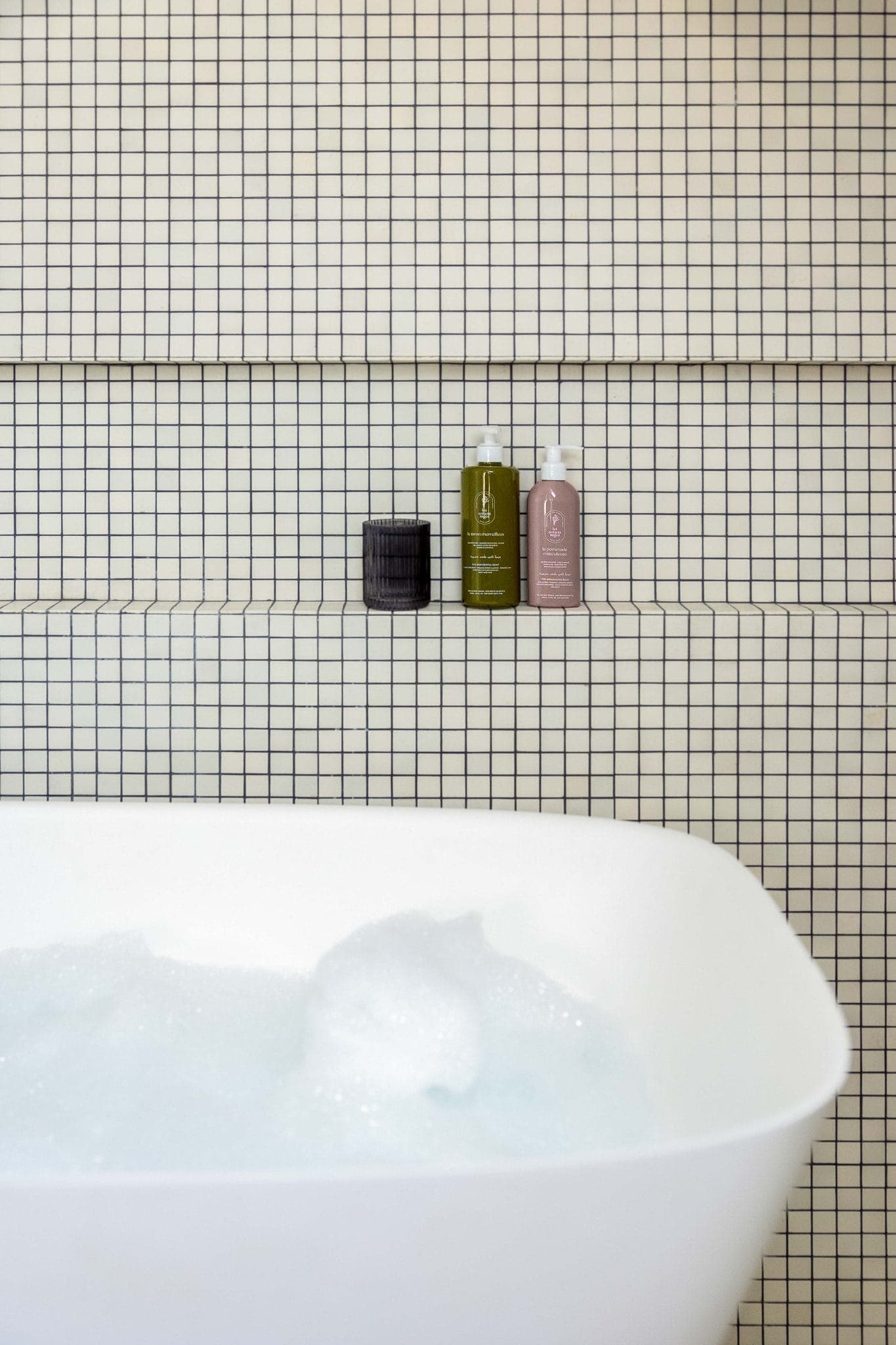 Au premier plan, une baignoire blanche remplie de bulles. Derrière, trois bouteilles et un petit récipient noir sont posés sur une étagère encastrée contre un mur recouvert de petits carreaux blancs carrés aux joints foncés.