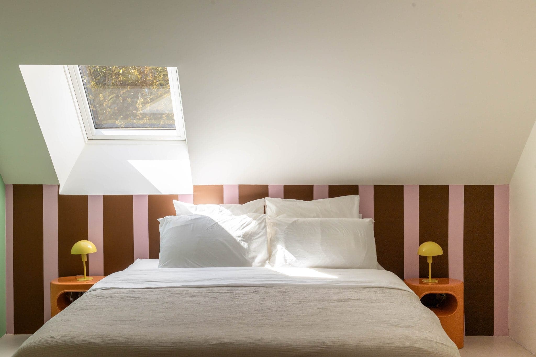 Un lit soigneusement fait avec une literie blanche trône sous un plafond mansardé percé d'une lucarne. La tête de lit est ornée de rayures verticales marron et roses, et de petites tables de chevet orange sont disposées de chaque côté, avec des lampes jaunes.