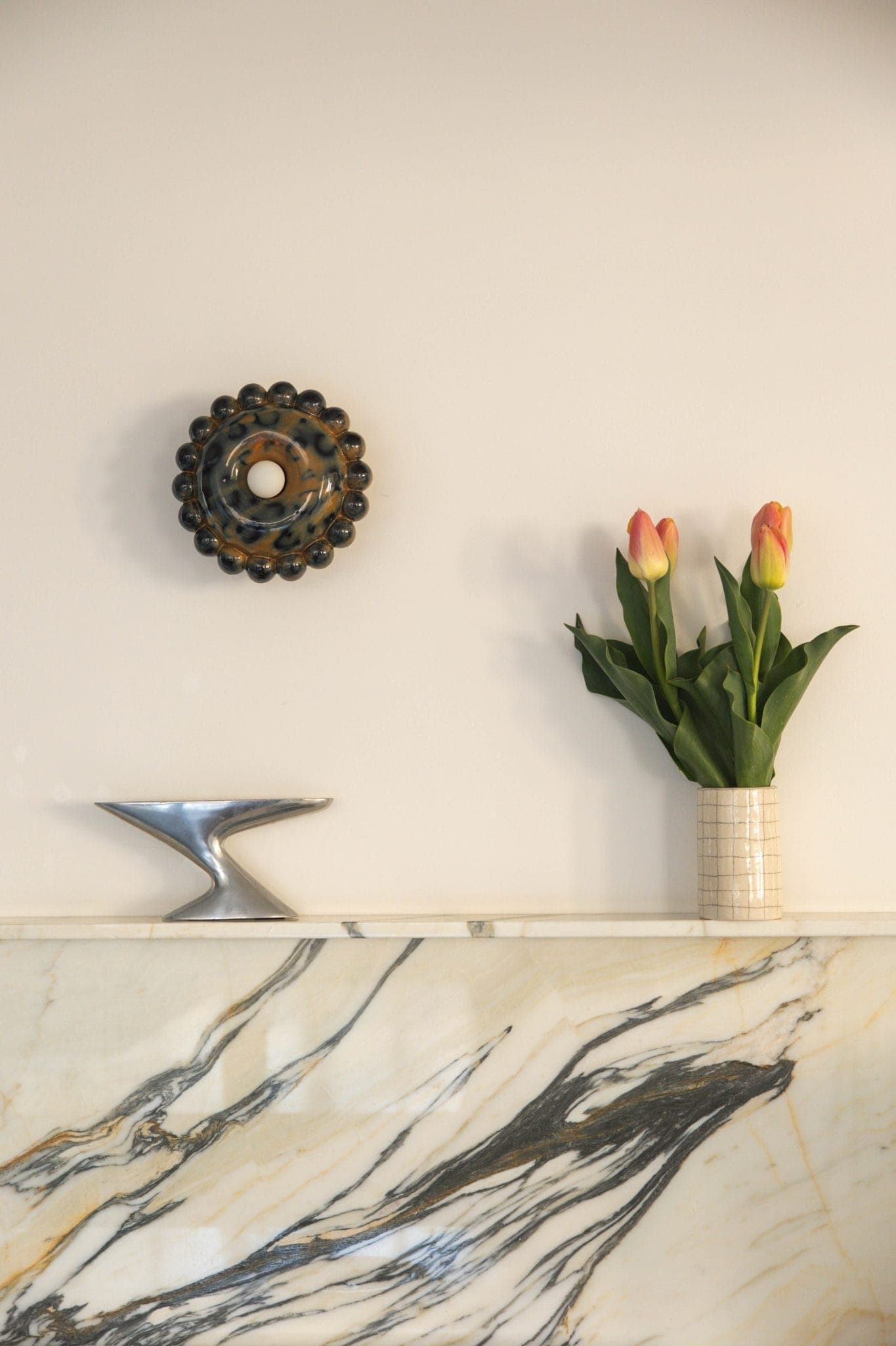 Une cheminée en marbre abrite une coupe décorative en argent et un vase contenant deux tulipes roses et jaunes. Au-dessus, une décoration murale ronde noire et dorée est accrochée sur un mur blanc cassé uni.