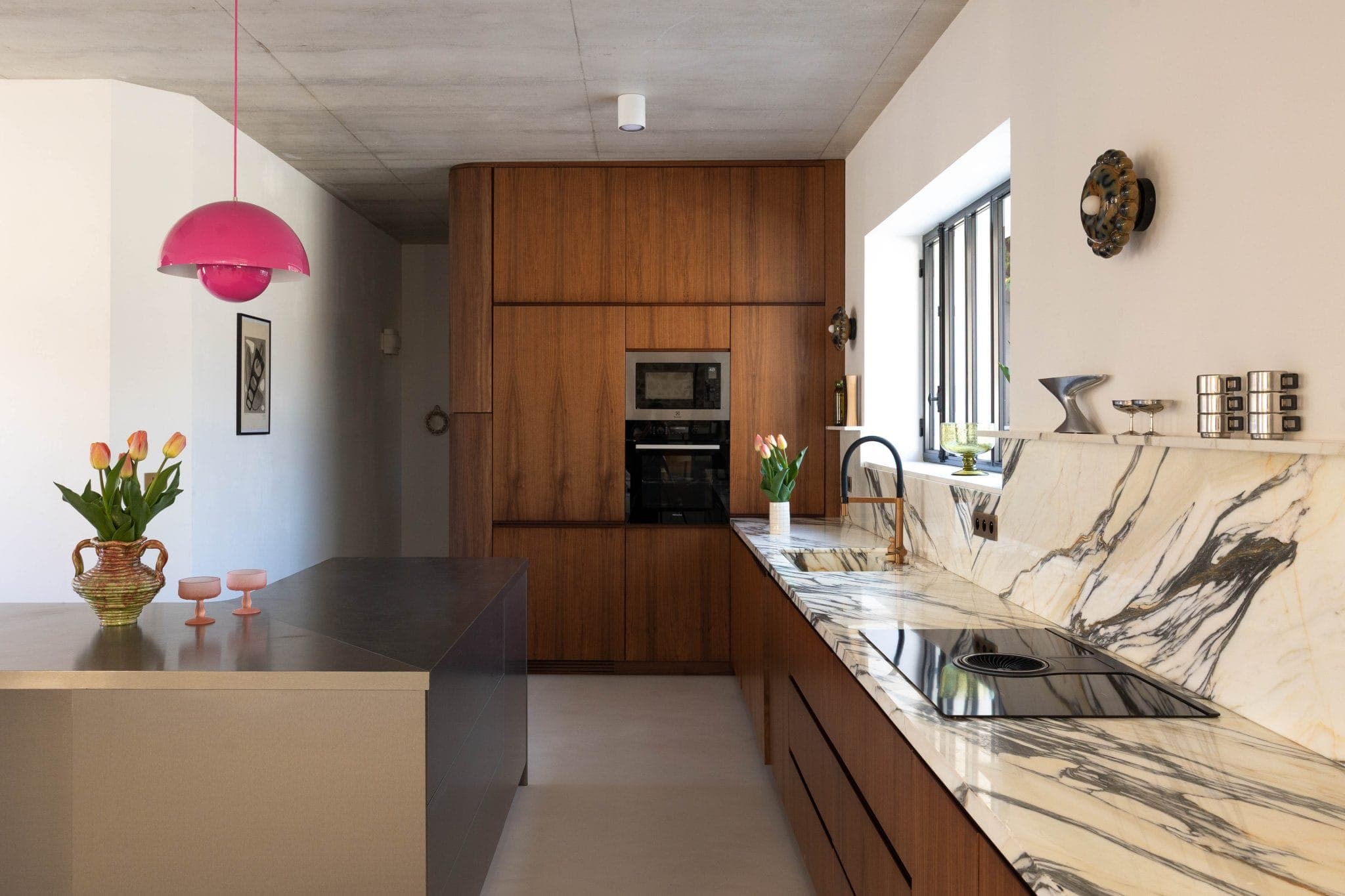 Cuisine moderne avec armoires en bois, comptoirs en marbre, îlot en acier inoxydable, suspension rose, vases de tulipes et une grande fenêtre laissant entrer la lumière naturelle.