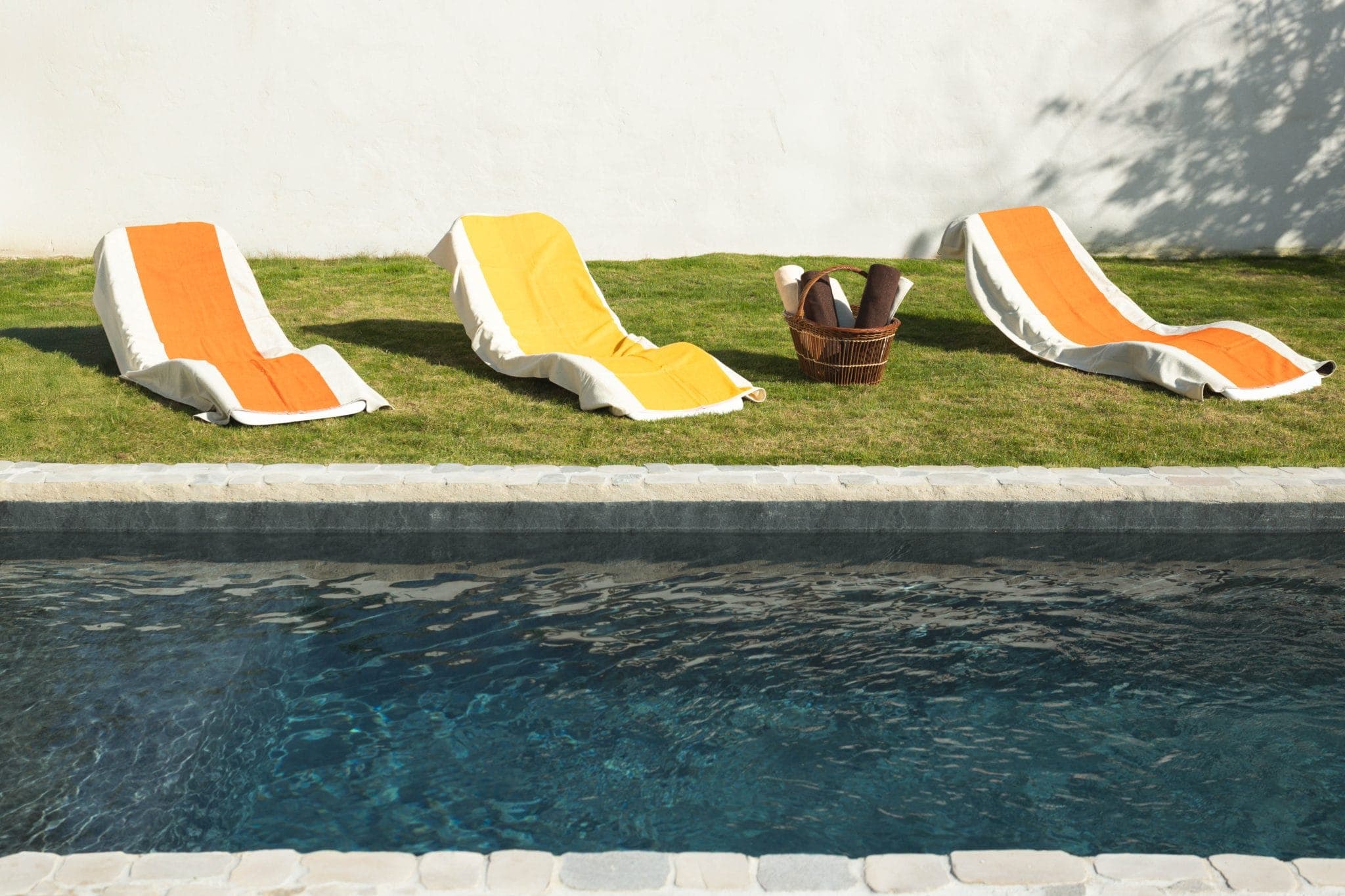 Trois chaises longues avec des serviettes orange et jaunes sont disposées sur l'herbe au bord d'une piscine. Un panier rempli de serviettes roulées est posé entre les chaises. La scène est ensoleillée, avec des ombres sur le mur.