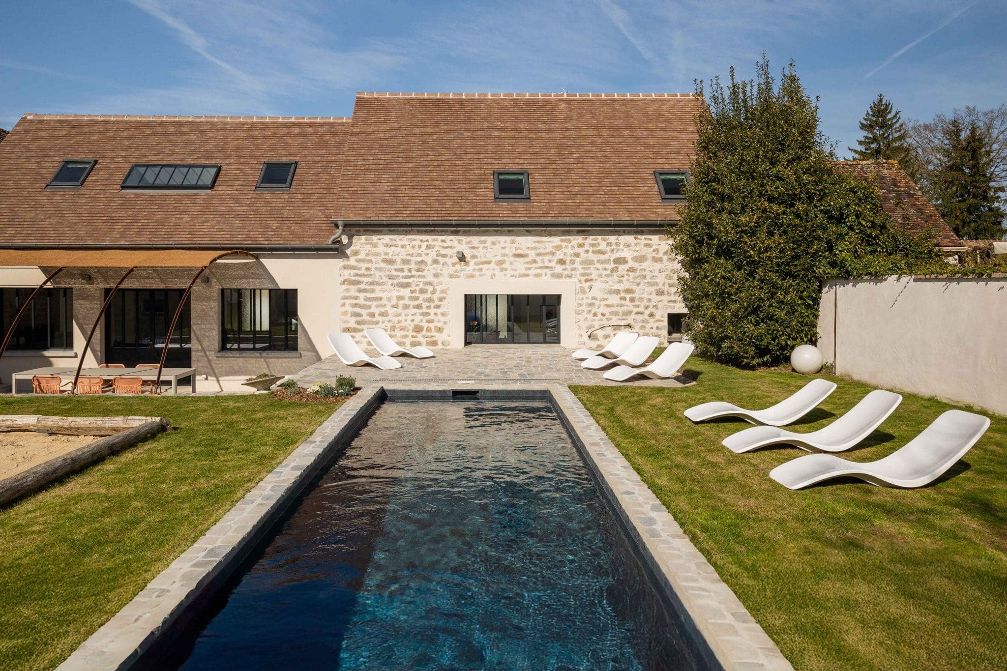 Une maison moderne à la façade en pierre dispose d'une longue piscine rectangulaire, entourée d'une pelouse verdoyante et de plusieurs chaises longues blanches. Le ciel est dégagé et l'espace extérieur est accueillant et serein.