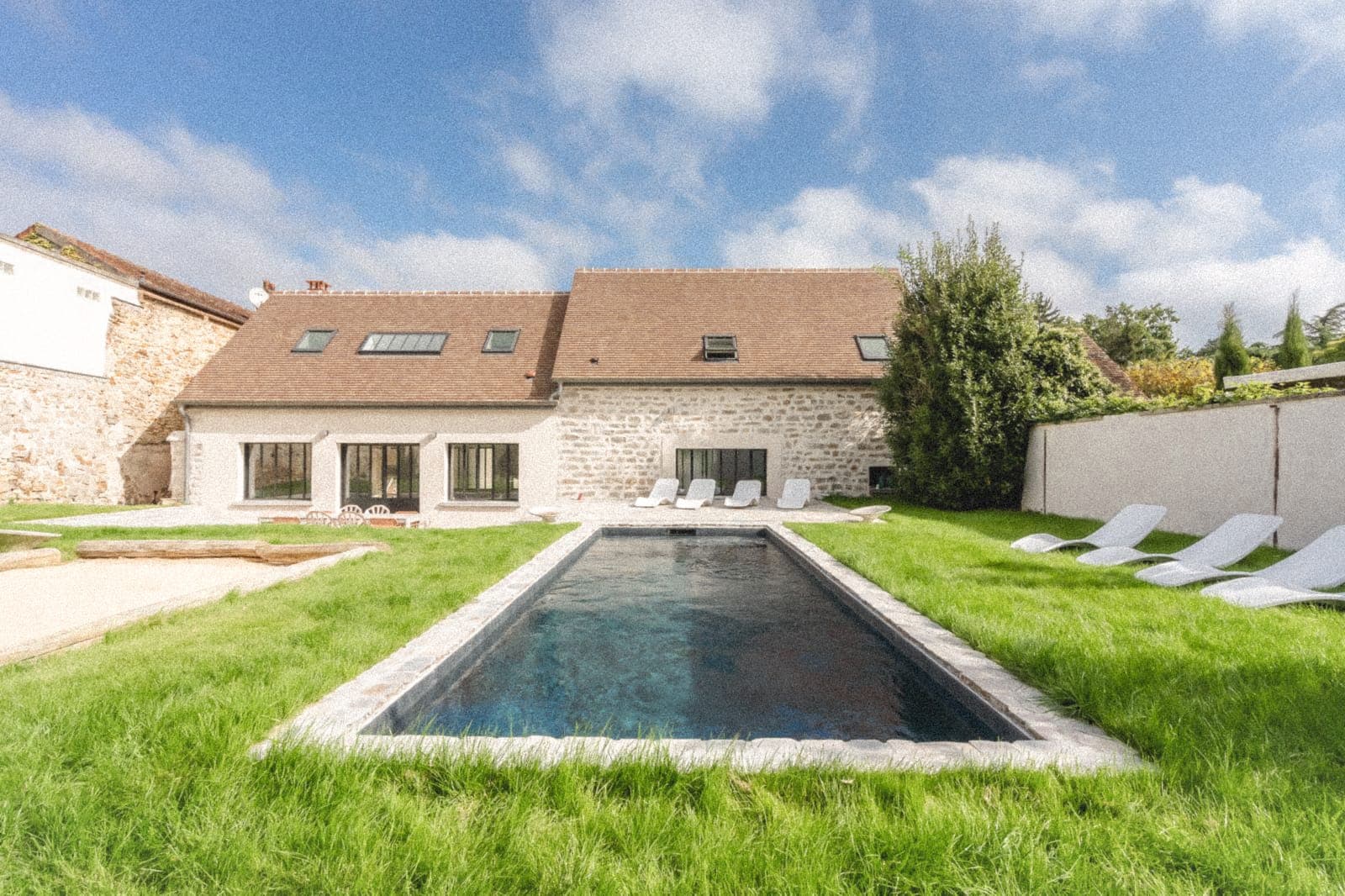 Maison en pierre au toit brun avec de multiples lucarnes, entourée d'herbes luxuriantes et d'arbres. Une longue et étroite piscine est au premier plan avec des chaises longues blanches disposées sur les côtés sous un ciel partiellement nuageux.