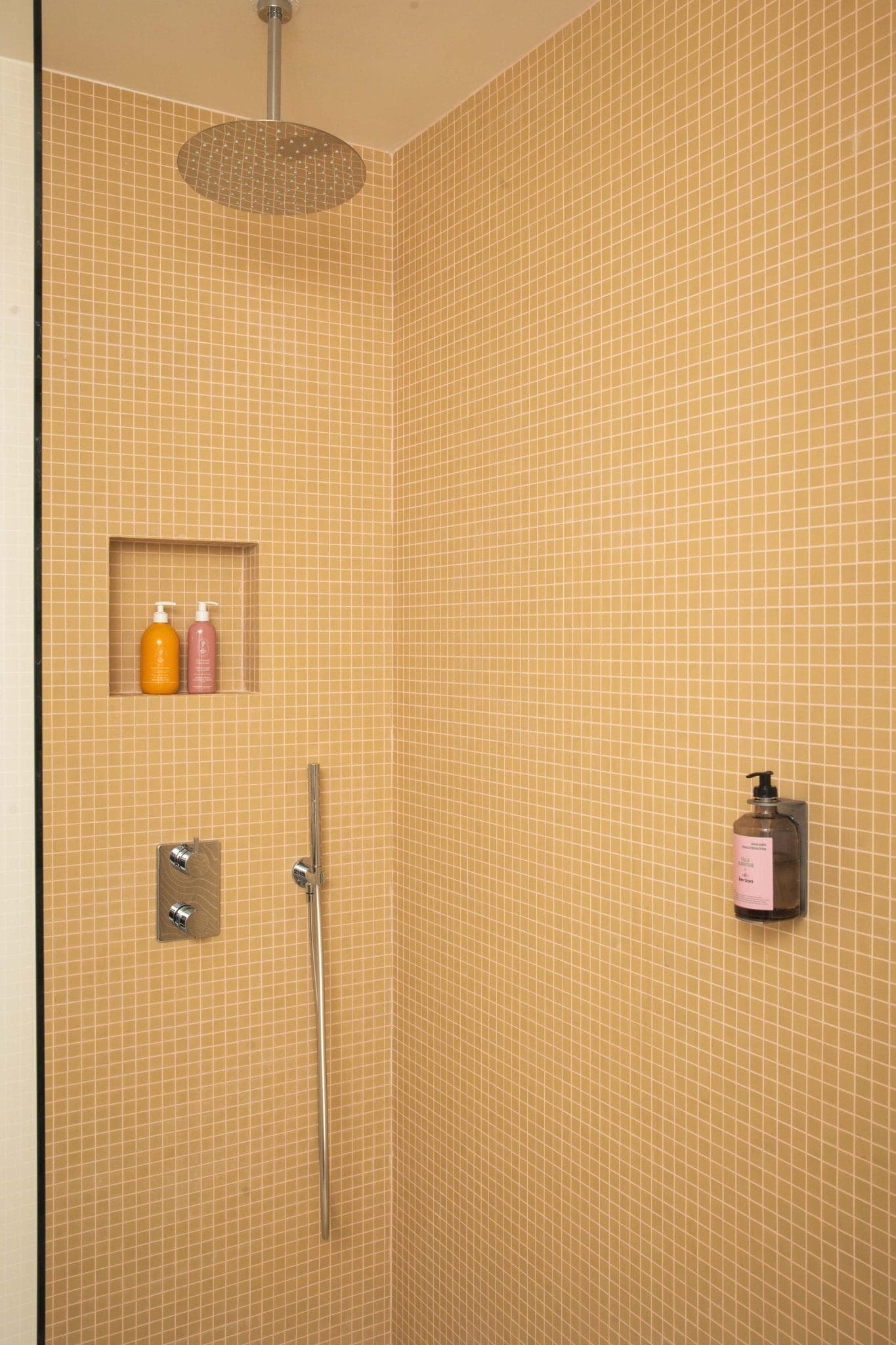 Une douche moderne avec de petits carreaux carrés orange-beige, une pomme de douche à effet pluie, deux étagères intégrées pour bouteilles, une barre de douche verticale et un distributeur de savon mural.