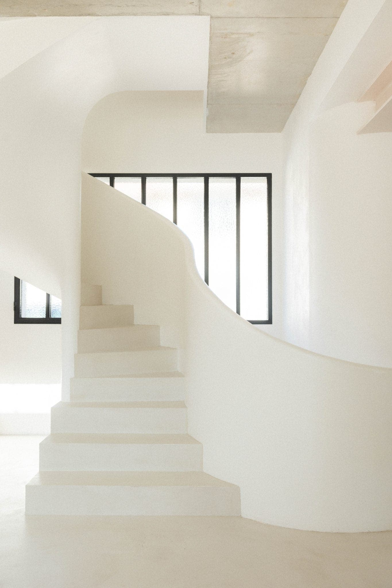 Escalier en colimaçon blanc minimaliste aux murs lisses et incurvés, menant à une pièce lumineuse. Une grande fenêtre rectangulaire aux cadres noirs laisse entrer la lumière naturelle, soulignant le design épuré et moderne.