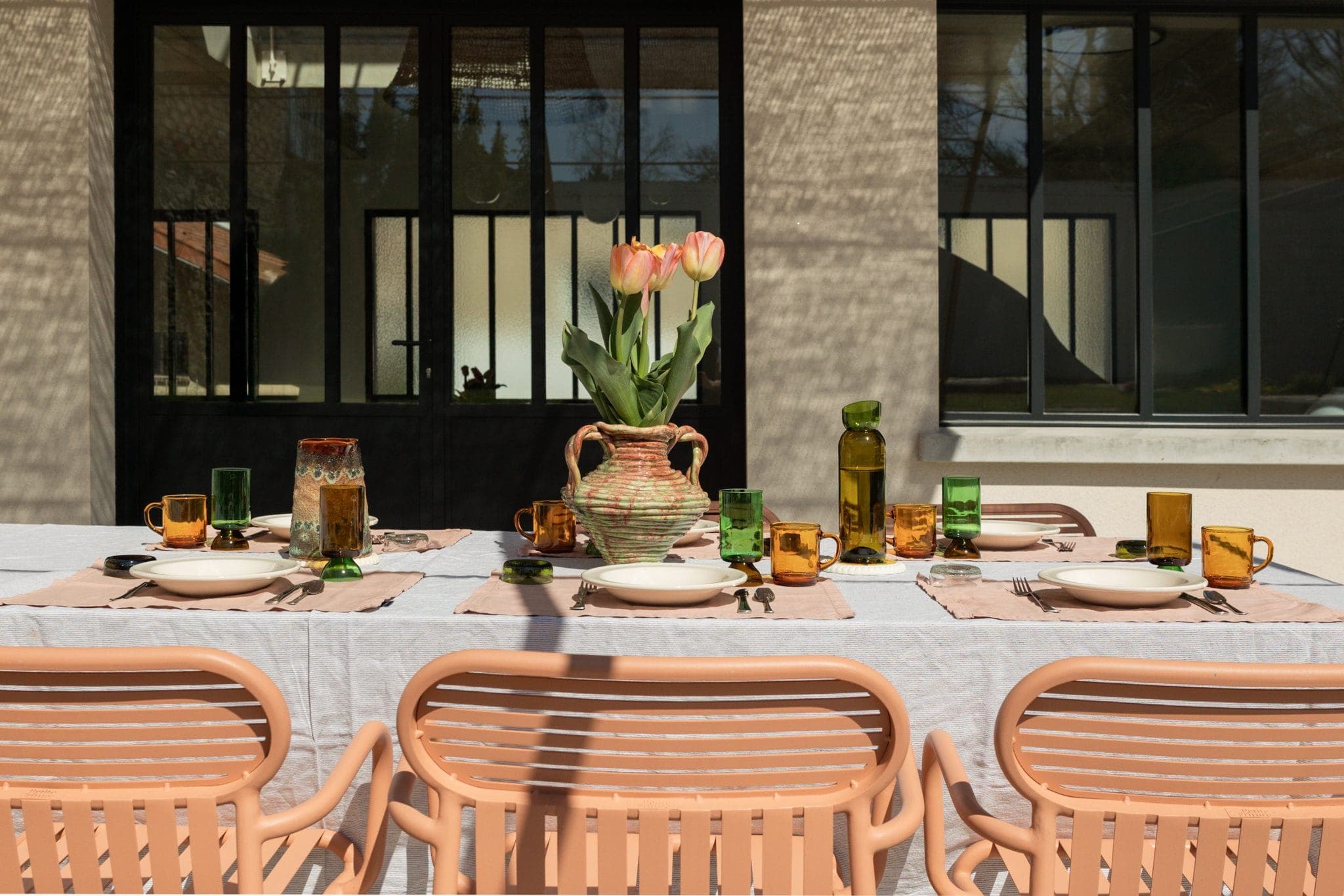 Une table à manger extérieure baignée de soleil est dressée avec des assiettes, des verres et un grand vase de tulipes roses. Quatre chaises couleur pêche sont disposées autour de la table, recouverte d'une nappe blanche et de sets de table beiges.