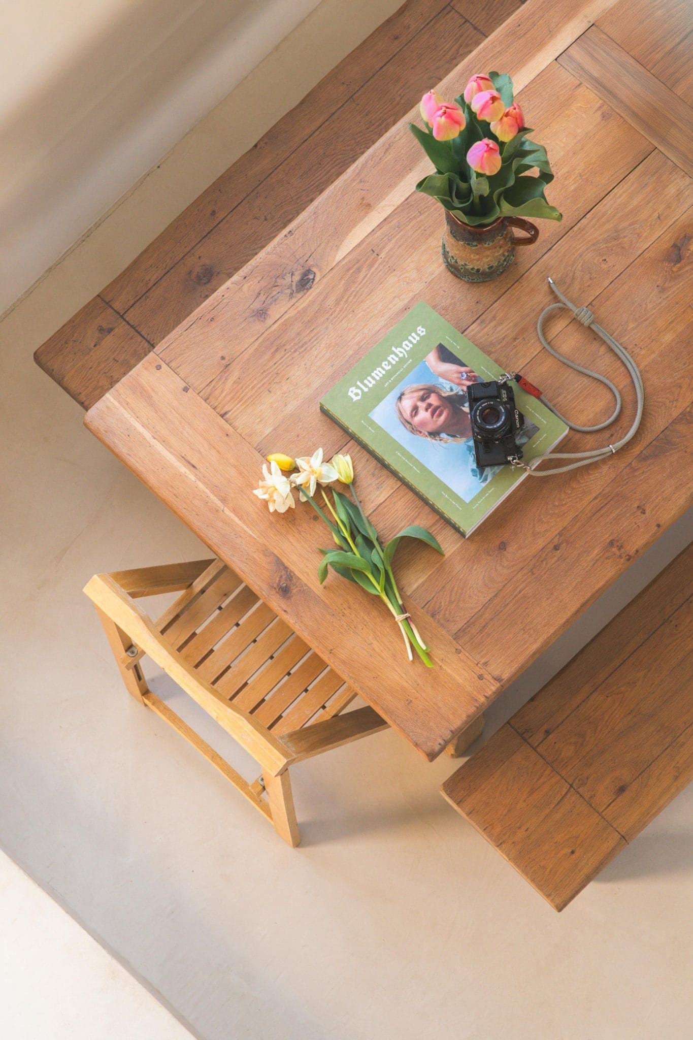 Une table en bois avec une chaise en bois, un vase de tulipes roses, un magazine vert avec le visage d'une femme sur la couverture, un appareil photo avec une sangle et un petit bouquet de fleurs blanches et jaunes sur le dessus.