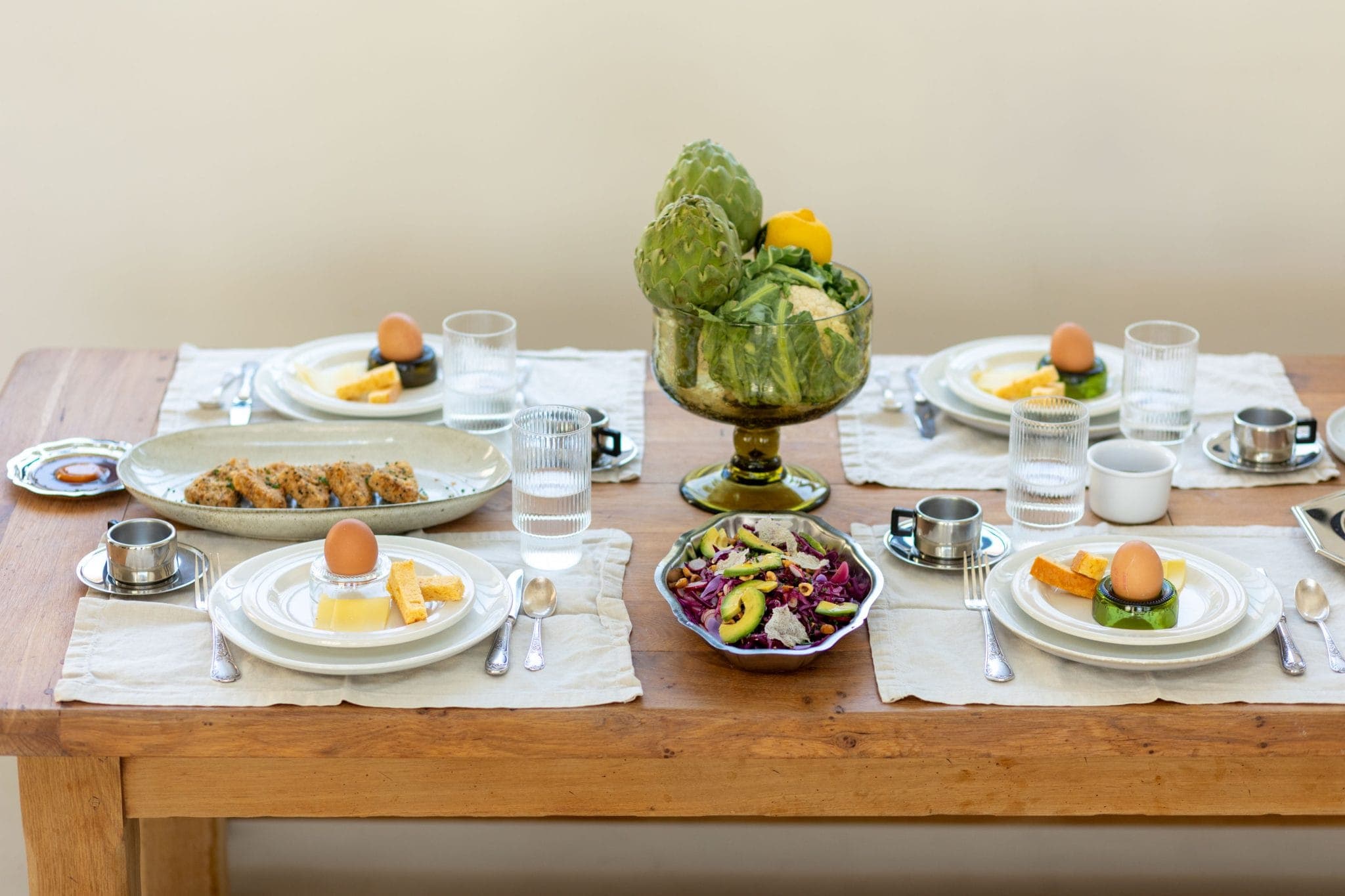 Une table à manger en bois dressée pour quatre personnes avec des assiettes d'œufs, de fromage et de légumes, des verres d'eau, un saladier, un plateau de légumes farcis et une pièce maîtresse d'artichauts et de poivrons frais.