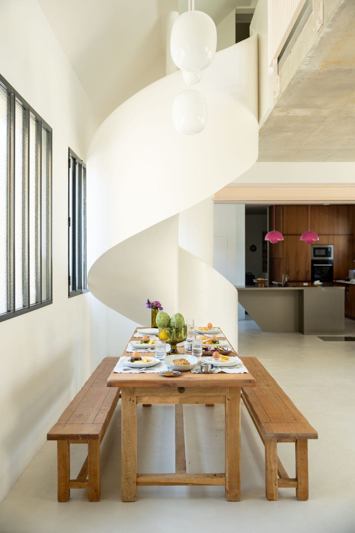 Une salle à manger lumineuse avec une longue table en bois dressée pour les repas, des bancs de chaque côté et un escalier en colimaçon blanc moderne en arrière-plan. De grandes fenêtres laissent entrer la lumière naturelle.