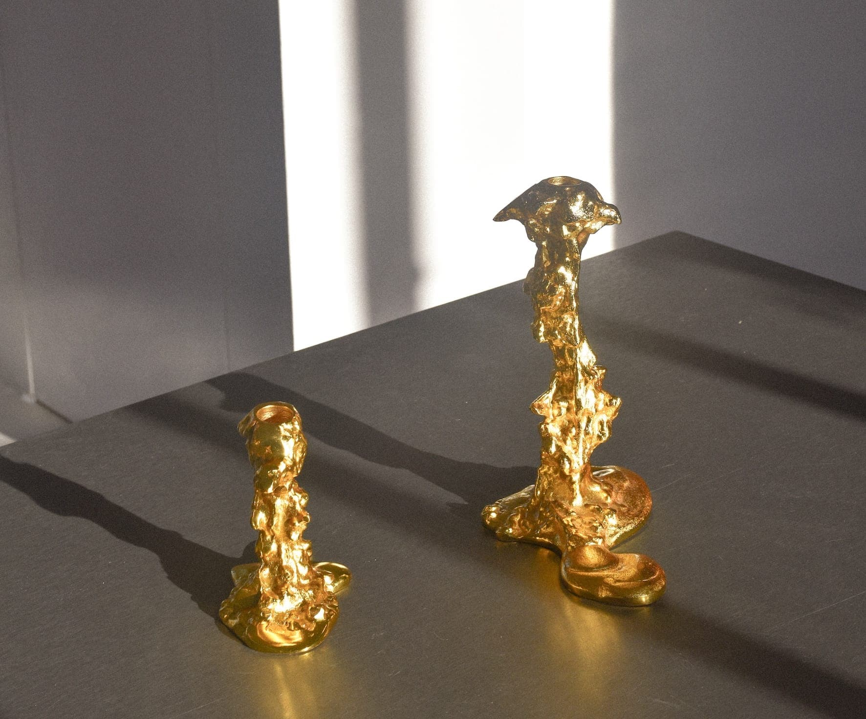 Deux sculptures abstraites dorées sur une surface sombre. Elles ont des formes irrégulières et allongées avec des textures organiques et sont placées à la lumière du soleil, projetant des ombres douces. L'arrière-plan est minimaliste, avec quelques lignes verticales dues à la lumière du soleil.