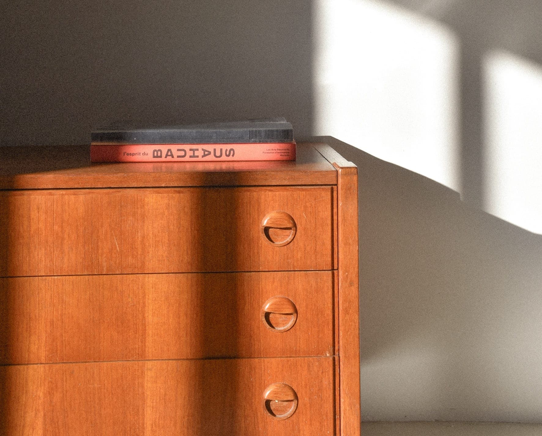 Une commode en bois à trois tiroirs est partiellement éclairée par la lumière du soleil. Un gros livre à la couverture rouge et au dos noir repose sur le dessus de la commode. Le mur derrière est peint en blanc, ce qui projette des ombres.