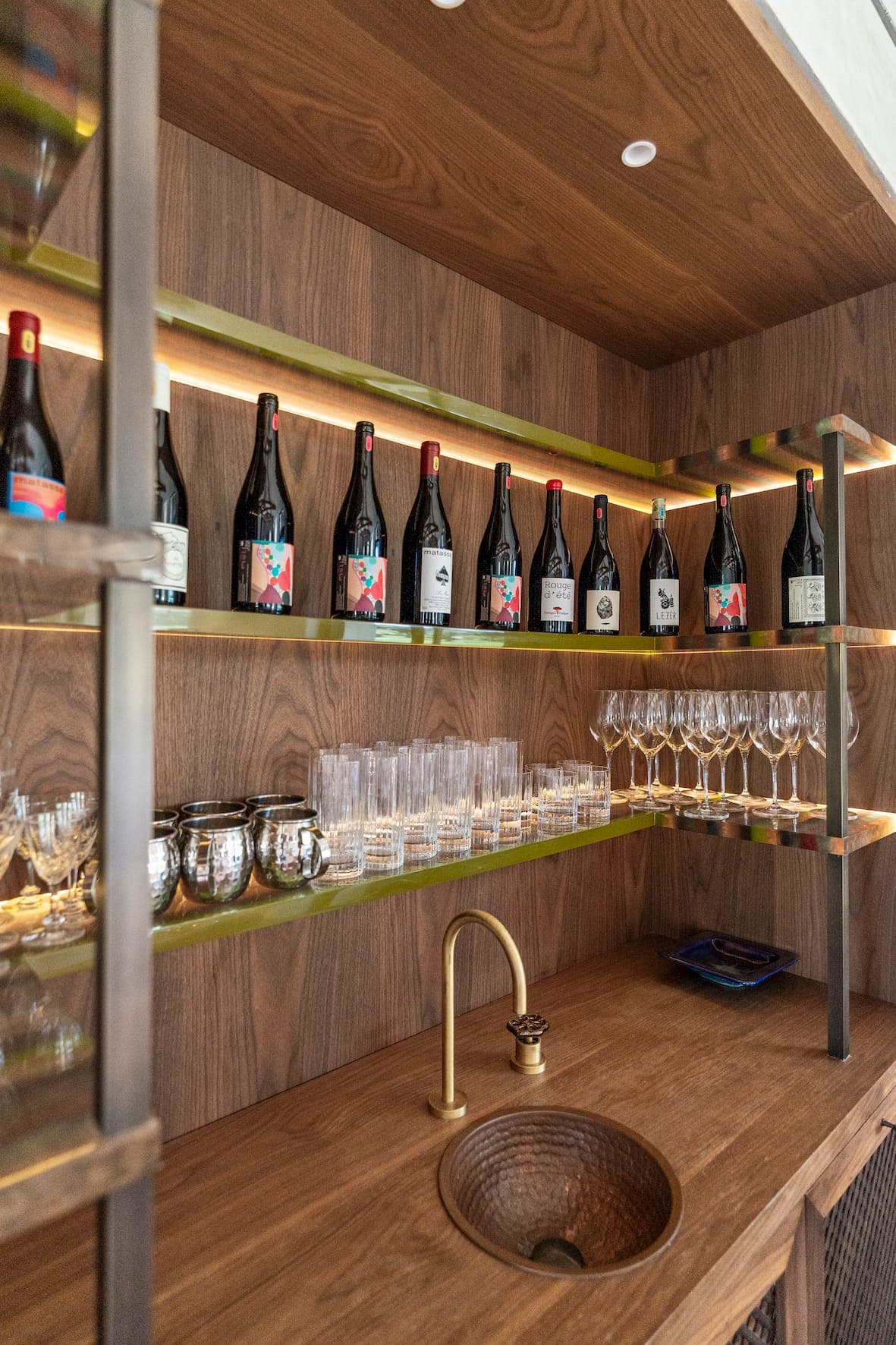 A modern wooden bar with shelves displaying various glasses and wine bottles. Several rows of wine bottles are arranged on the upper shelves, with glasses underneath. A sink with brass faucet is integrated into the worktop, accompanied by metal bowls.