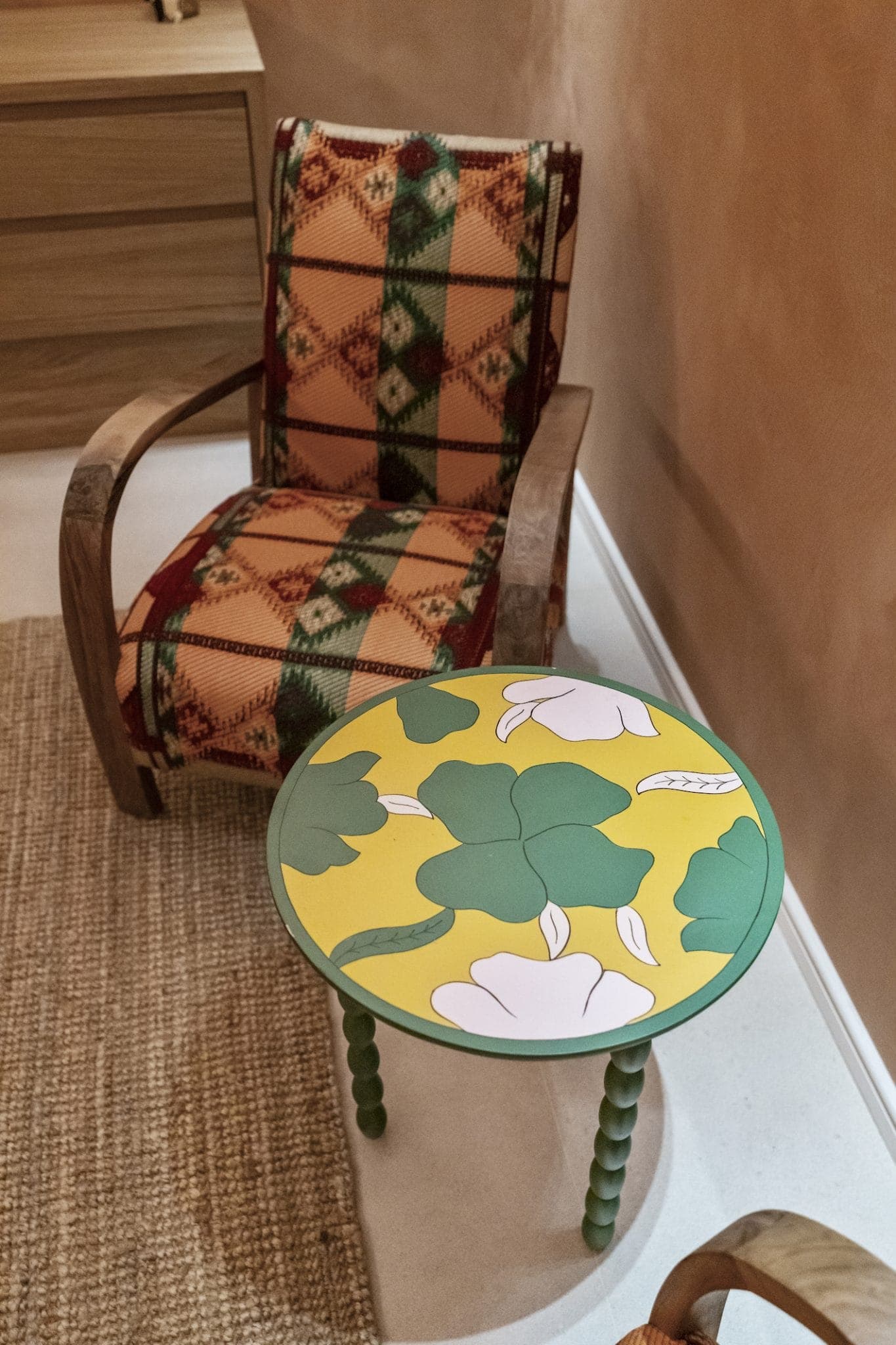 A wooden chair covered in multicolored tartan fabric sits next to a small round table. The table has a bright yellow top with green leaves and white flowers. A natural-fiber rug is visible on the floor. The walls are beige.
