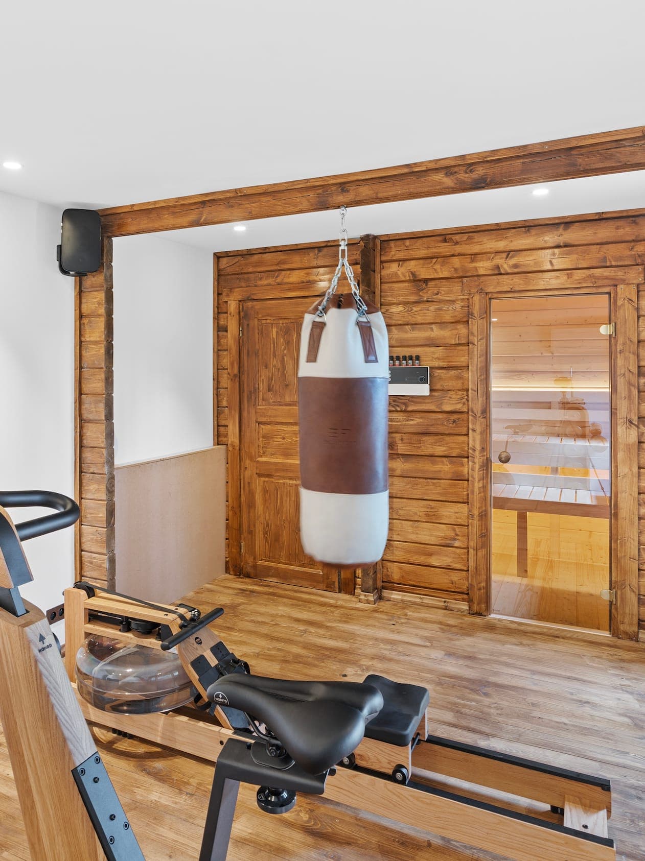 Wooden-walled and wood-floored gym with hanging punching bag, rowing machine and stationary bike. A sauna with glass door is in the background. Bright lighting illuminates the space.