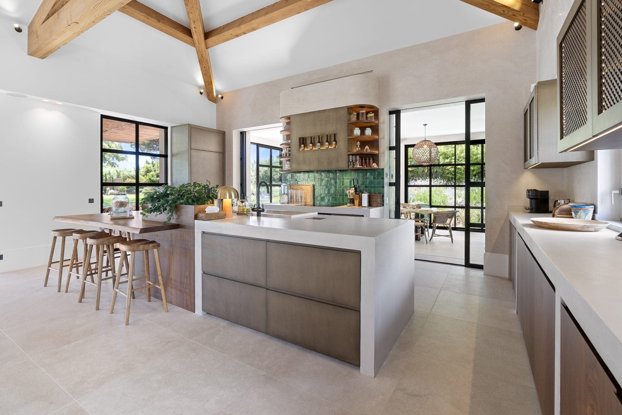 Spacious, modern kitchen with large central island, wooden stools and exposed beams. The light-colored floor complements the earthy tones. Green tiles and large windows add contrast and light to the room.