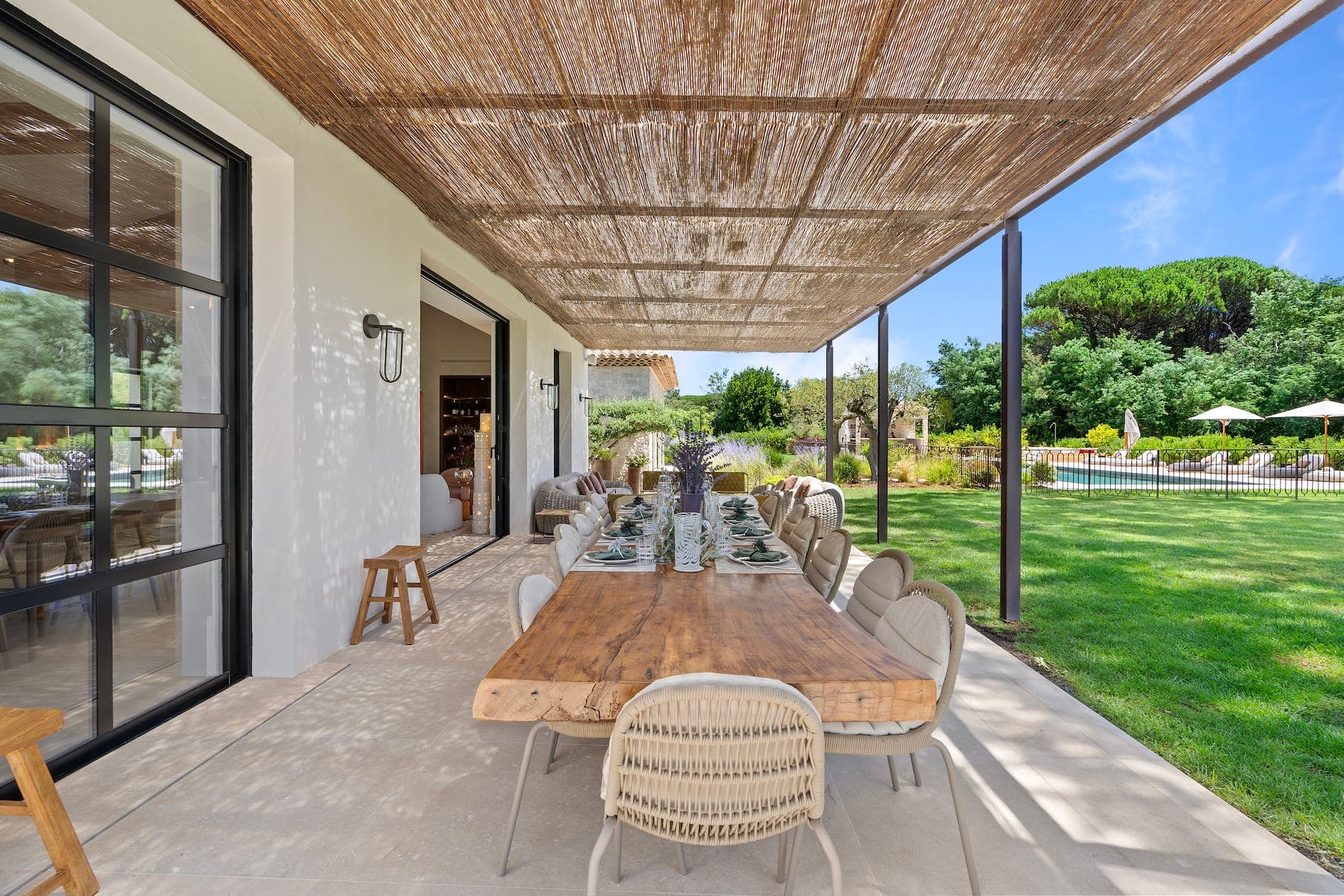 Outdoor terrace with long wooden dining table. A woven awning provides shade. The area is surrounded by a well-manicured lawn and large trees, with a swimming pool and sun loungers visible in the background.