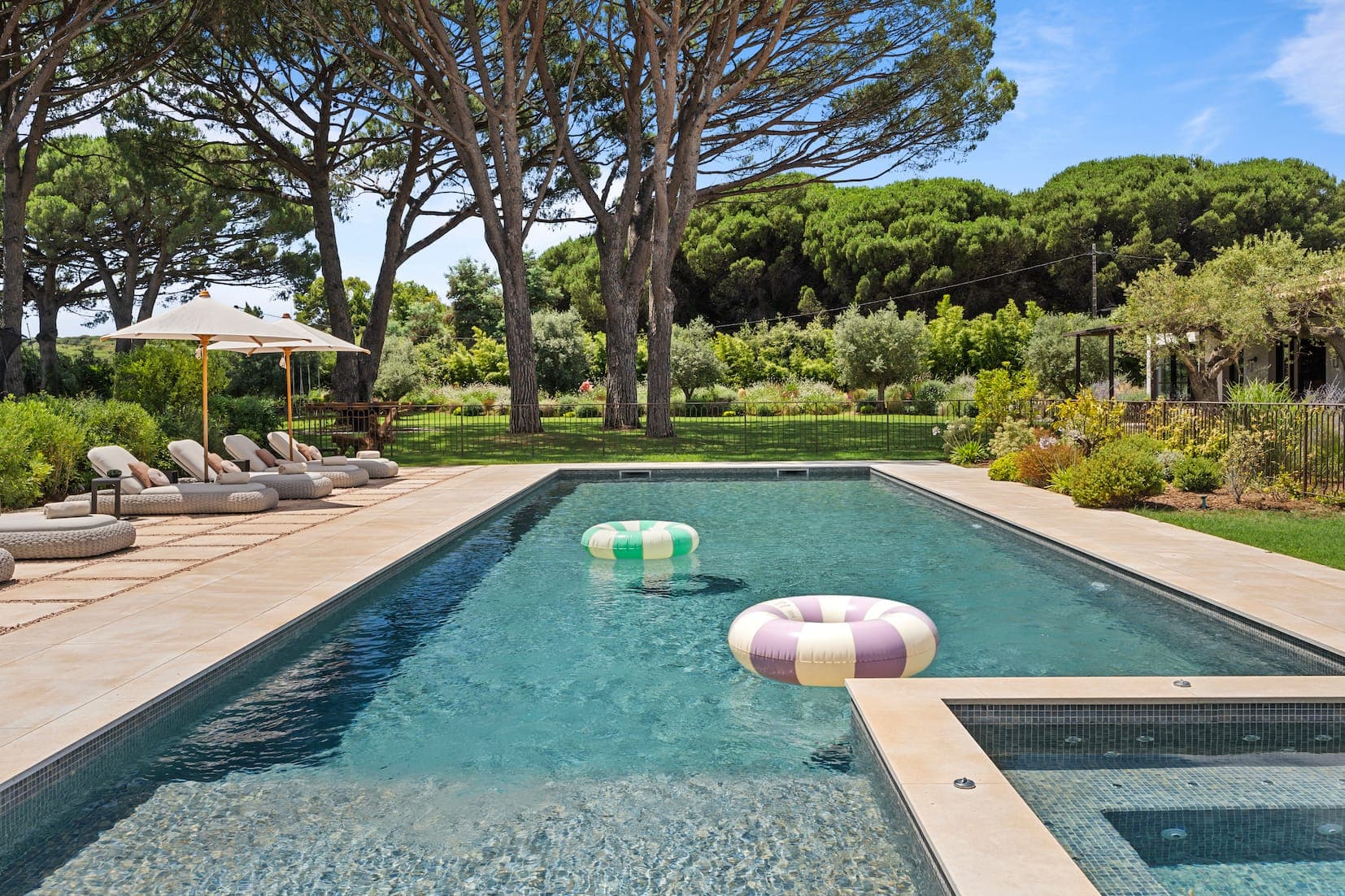 A rectangular swimming pool with two inflatable rings, surrounded by lounge chairs and umbrellas. The area is bordered by lush greenery and tall trees, with a clear blue sky overhead.