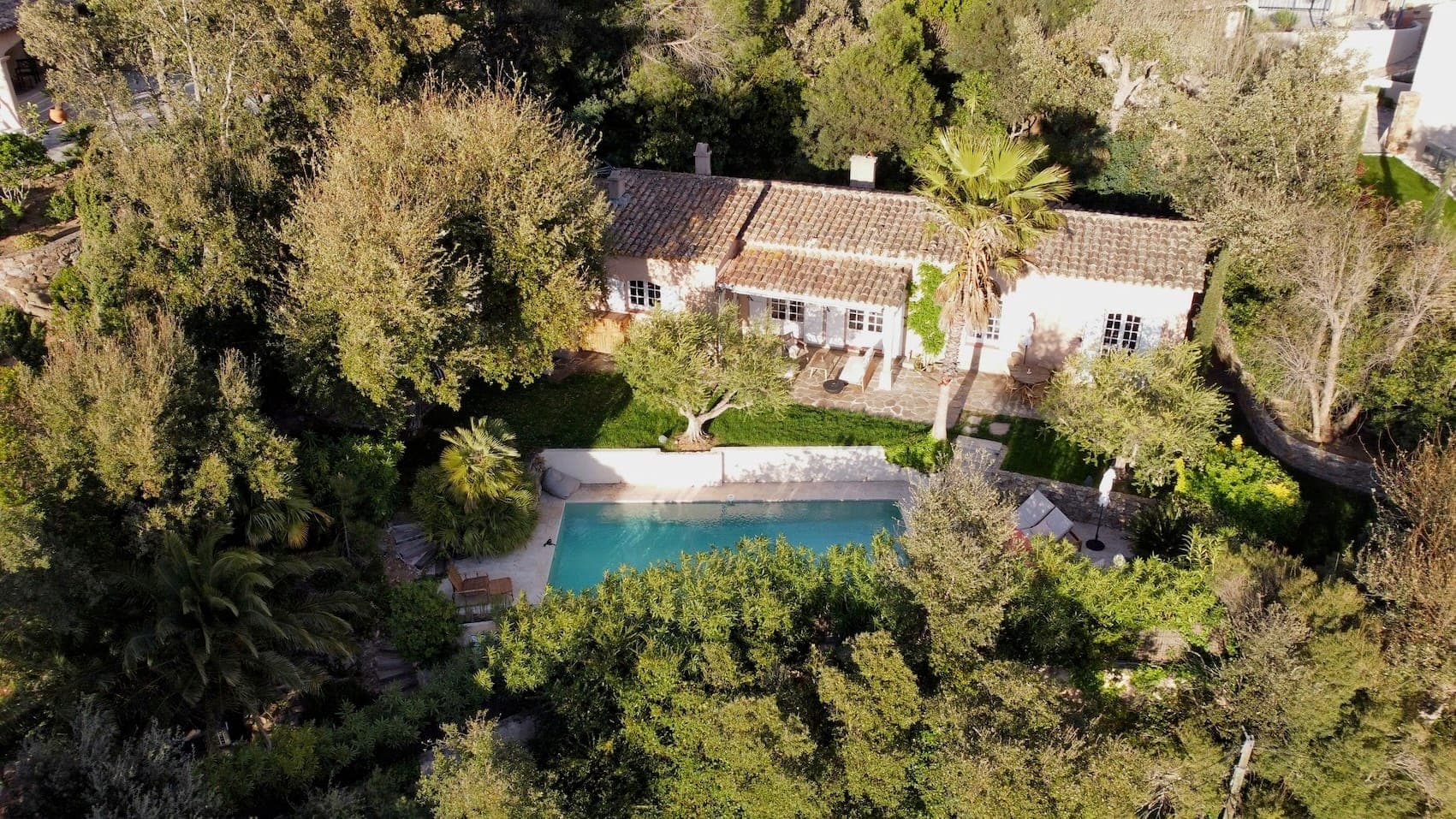 Aerial view of a charming house surrounded by dense greenery, featuring a lush garden with a swimming pool. The house has a terracotta roof and white walls, nestled amid tall trees and a variety of plants.