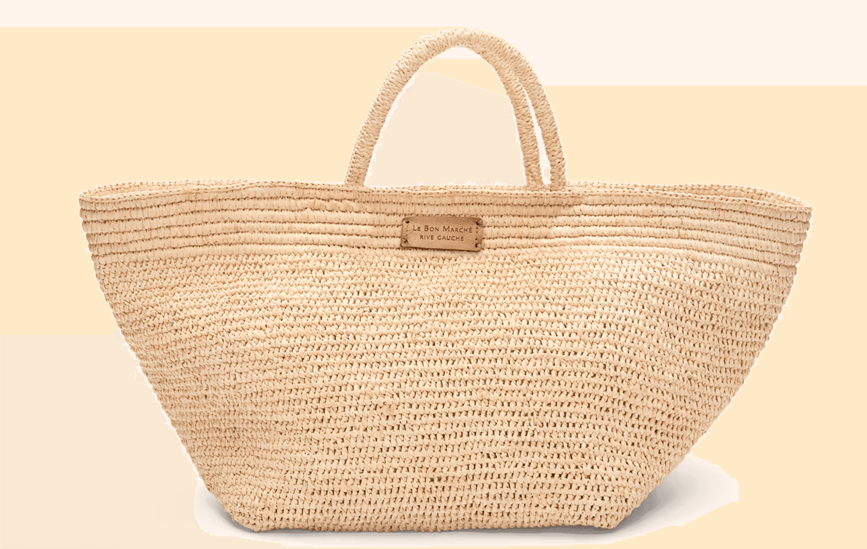 Un grand sac fourre-tout en paille tressée avec deux anses et un petit empiècement en cuir affichant un texte sur le devant. Le sac se présente sur un fond beige uni.