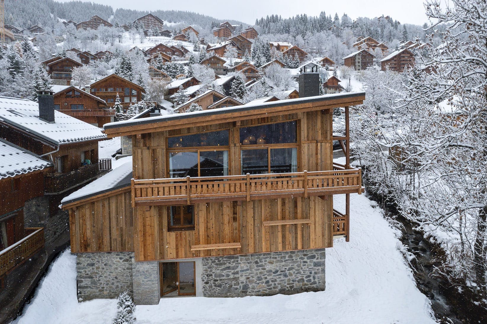 A wooden chalet with large windows and balconies is covered in snow, on a snow-covered hillside with other chalets scattered in the background. The area is surrounded by trees and snow-covered roofs.