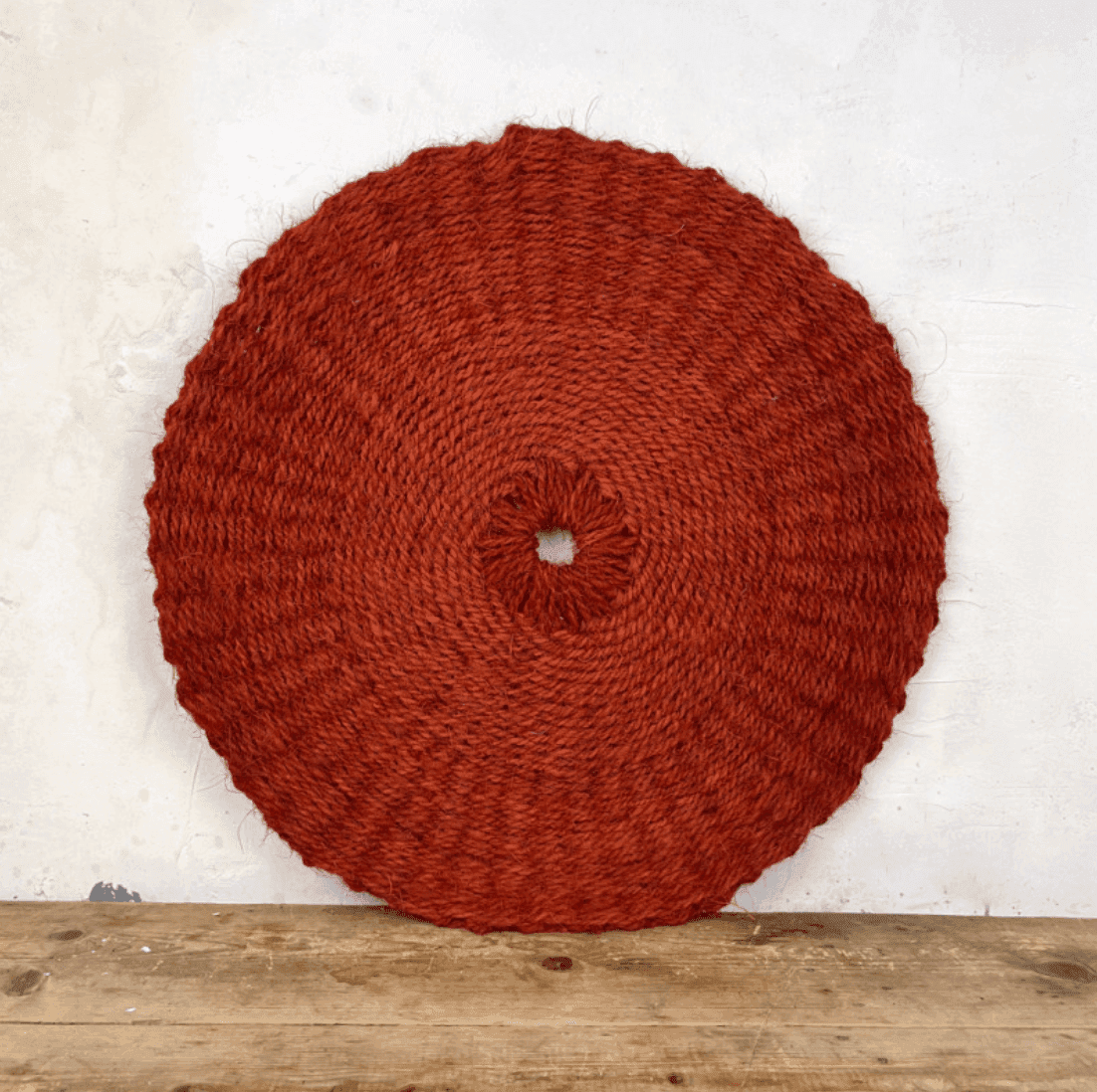 Un tapis rond tissé de couleur rouille avec un motif texturé est placé sur une surface en bois sur un fond gris clair. Un petit trou est visible au centre du tapis.